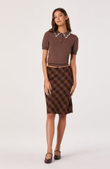 A woman is wearing a brown short-sleeved sweater with a collared neckline decorated with white floral embroidery along the edges. She pairs it with a brown plaid knee-length skirt with diagonal lines in shades of brown, yellow, and orange. She completes the outfit with matching brown shoes with ankle straps.