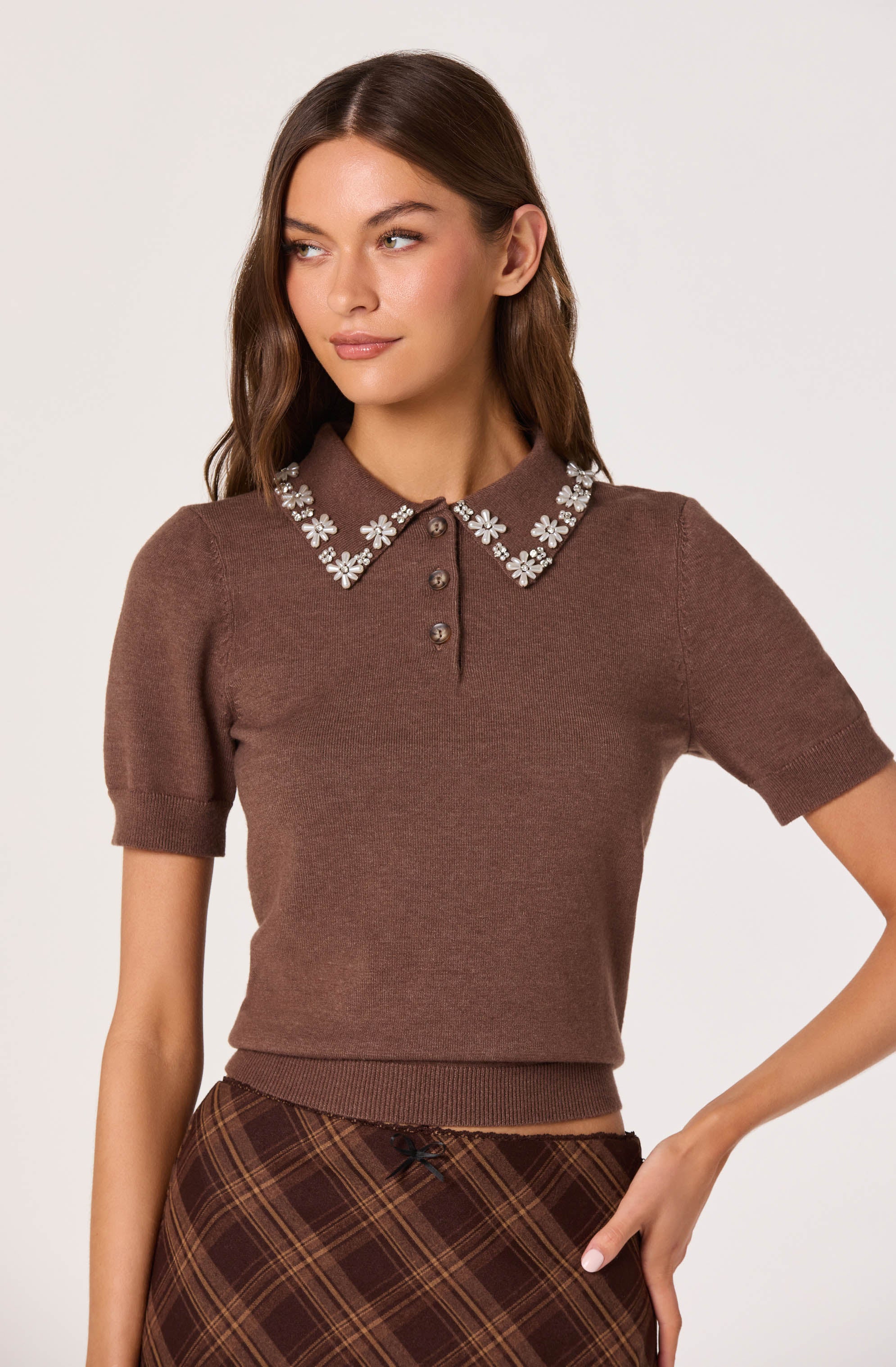 A woman is wearing a brown short-sleeved sweater with a ribbed waistband and cuffs. The sweater features a pointed collar decorated with white floral embroidery and has a button placket with four buttons. She is also wearing a brown plaid skirt with diagonal lines and a small bow at the waistband.