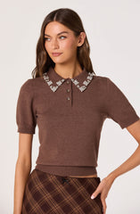 A woman is wearing a brown short-sleeved sweater with a ribbed waistband and cuffs. The sweater features a pointed collar decorated with white floral embroidery and has a button placket with four buttons. She is also wearing a brown plaid skirt with diagonal lines and a small bow at the waistband.