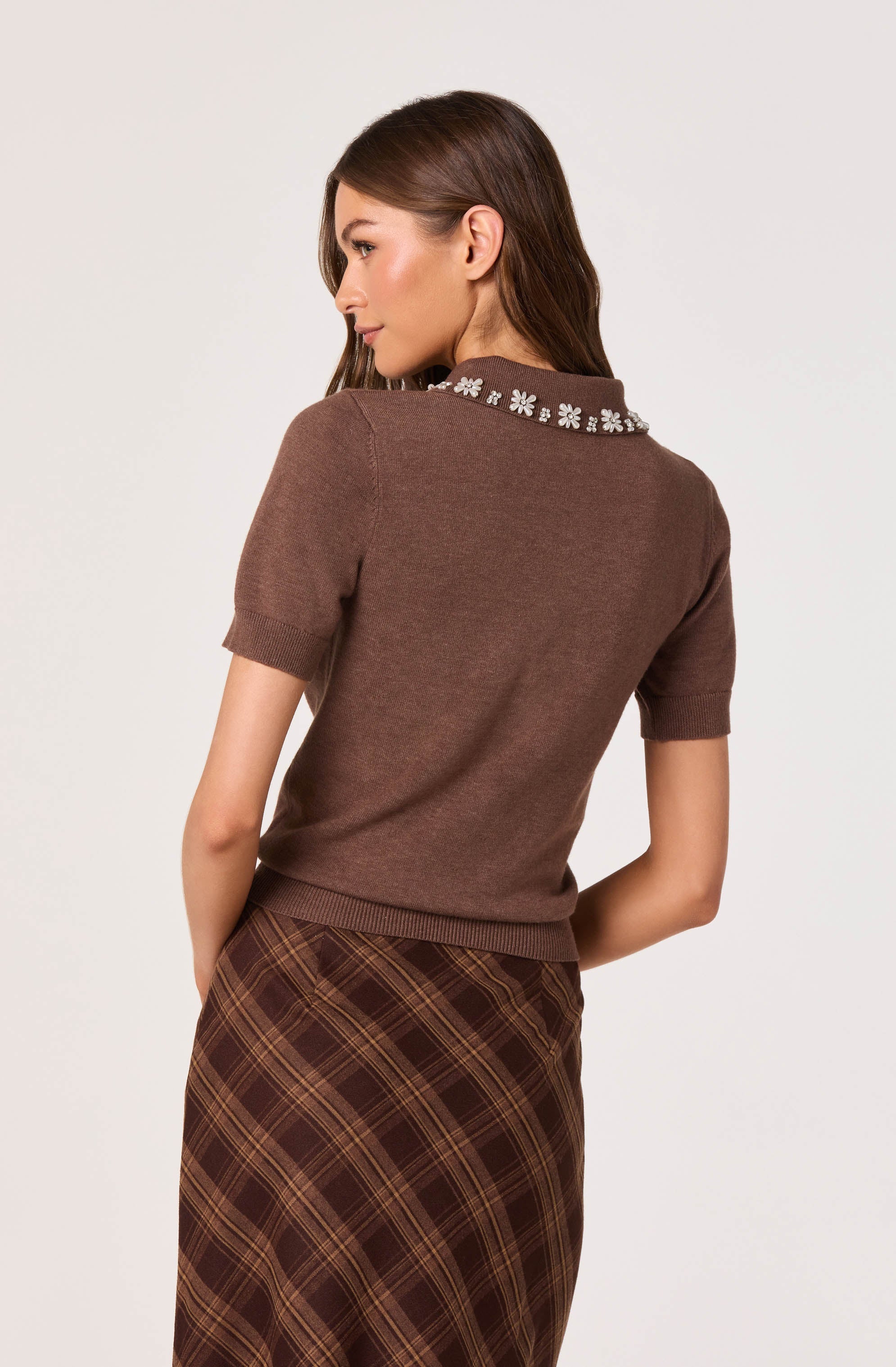 A woman wearing a short-sleeved brown sweater with a decorative collar adorned with white floral embroidery. She pairs it with a high-waisted, brown plaid skirt featuring diagonal stripes in shades of brown and yellow.