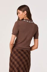 A woman wearing a short-sleeved brown sweater with a decorative collar adorned with white floral embroidery. She pairs it with a high-waisted, brown plaid skirt featuring diagonal stripes in shades of brown and yellow.