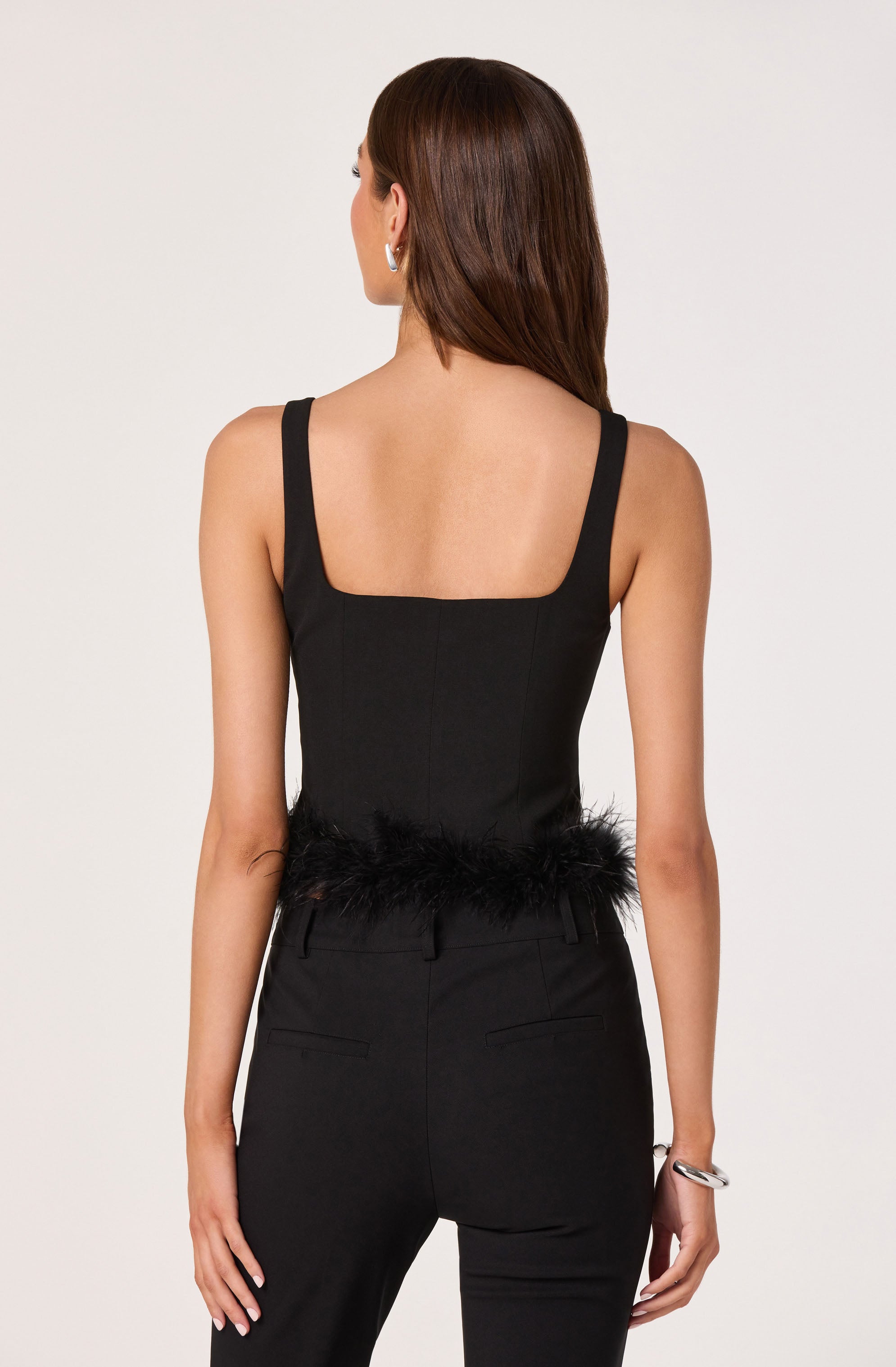 A woman is wearing a sleeveless black top with wide shoulder straps and a square neckline, paired with high-waisted black trousers. The top features a feathered trim at the waistline.