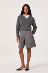 A woman is wearing a dark gray sweater with a V-neckline layered over a light gray collared shirt. She pairs this with high-waisted, knee-length gray shorts that have front pleats and a side seam detail. She completes her outfit with black pointed-toe high heels featuring a bow embellishment on the front.