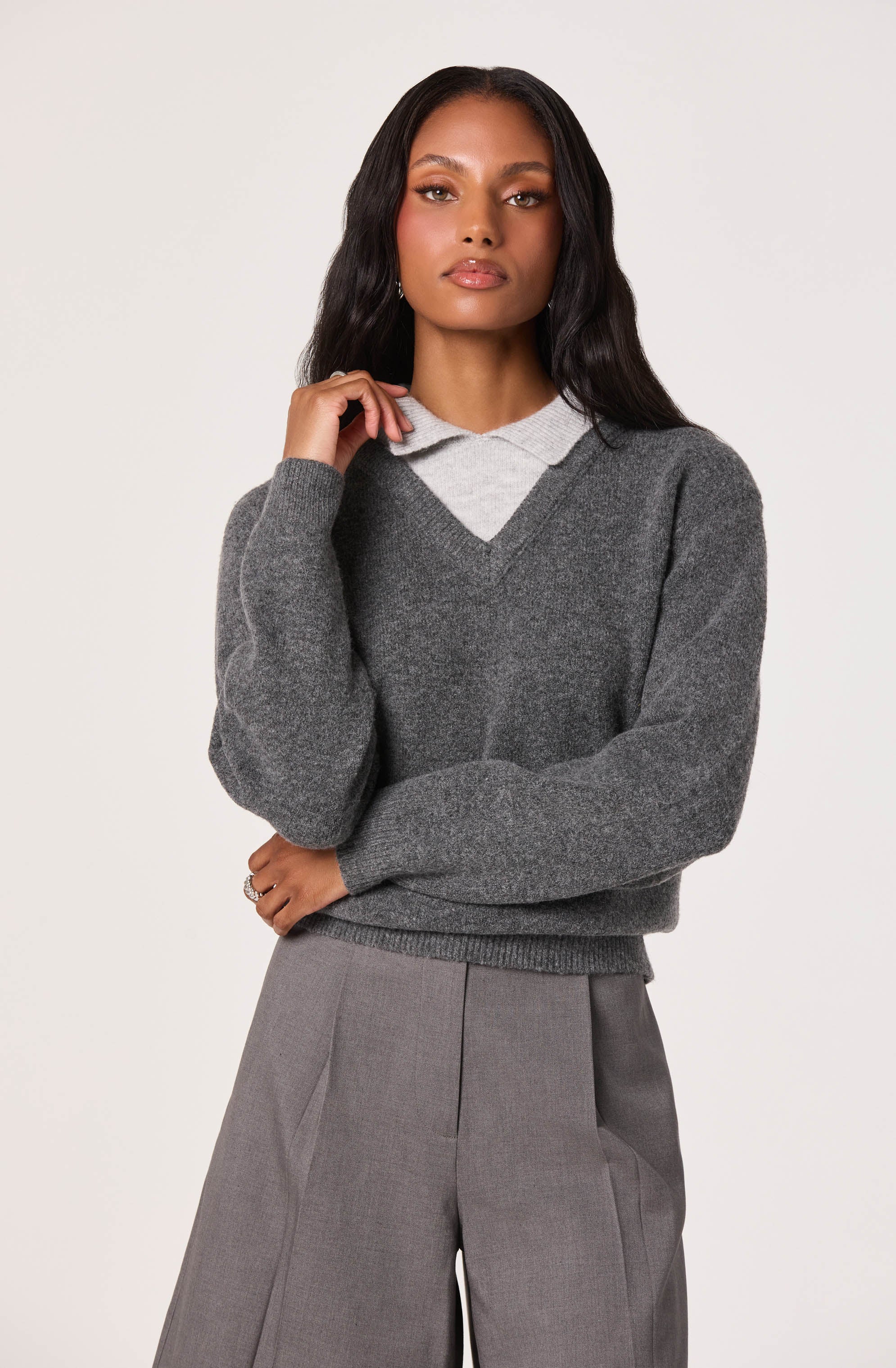 A woman is wearing a dark gray V-neck sweater layered over a light gray collared shirt, paired with high-waisted gray trousers.