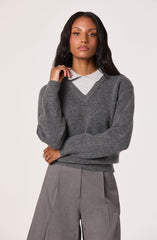 A woman is wearing a dark gray V-neck sweater layered over a light gray collared shirt, paired with high-waisted gray trousers.