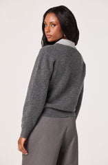 A woman is wearing a gray wool sweater with a light gray collar, paired with gray dress pants. The sweater has a ribbed hem and cuffs, and the overall look is elegant and professional. The woman has long, dark hair and is posed against a plain, light-colored background.