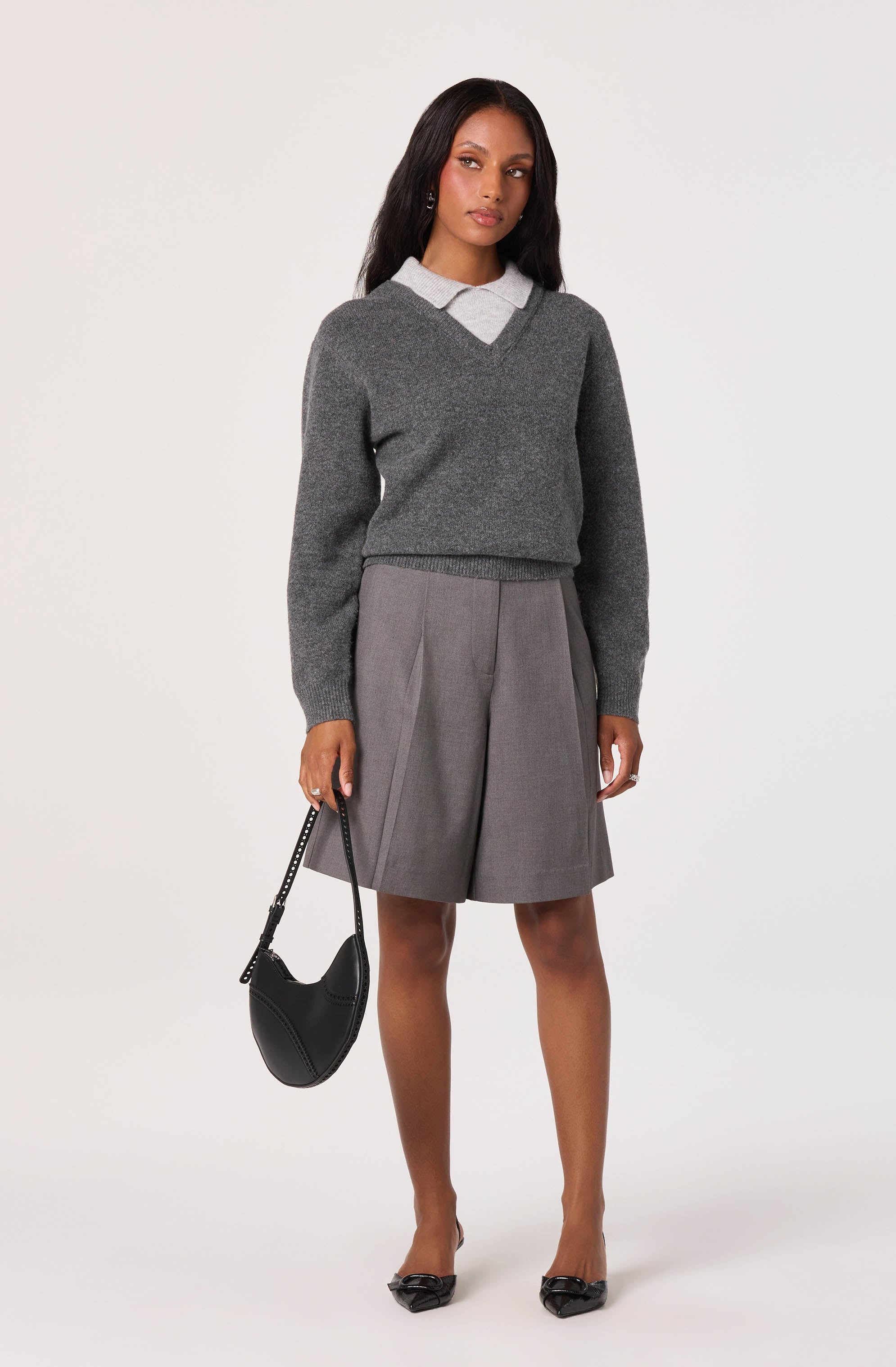 A woman dressed in a light gray collared shirt layered with a dark gray V-neck sweater, paired with knee-length gray pleated shorts. She holds a black handbag with a chain strap in her right hand and wears black pointed-toe heels.
