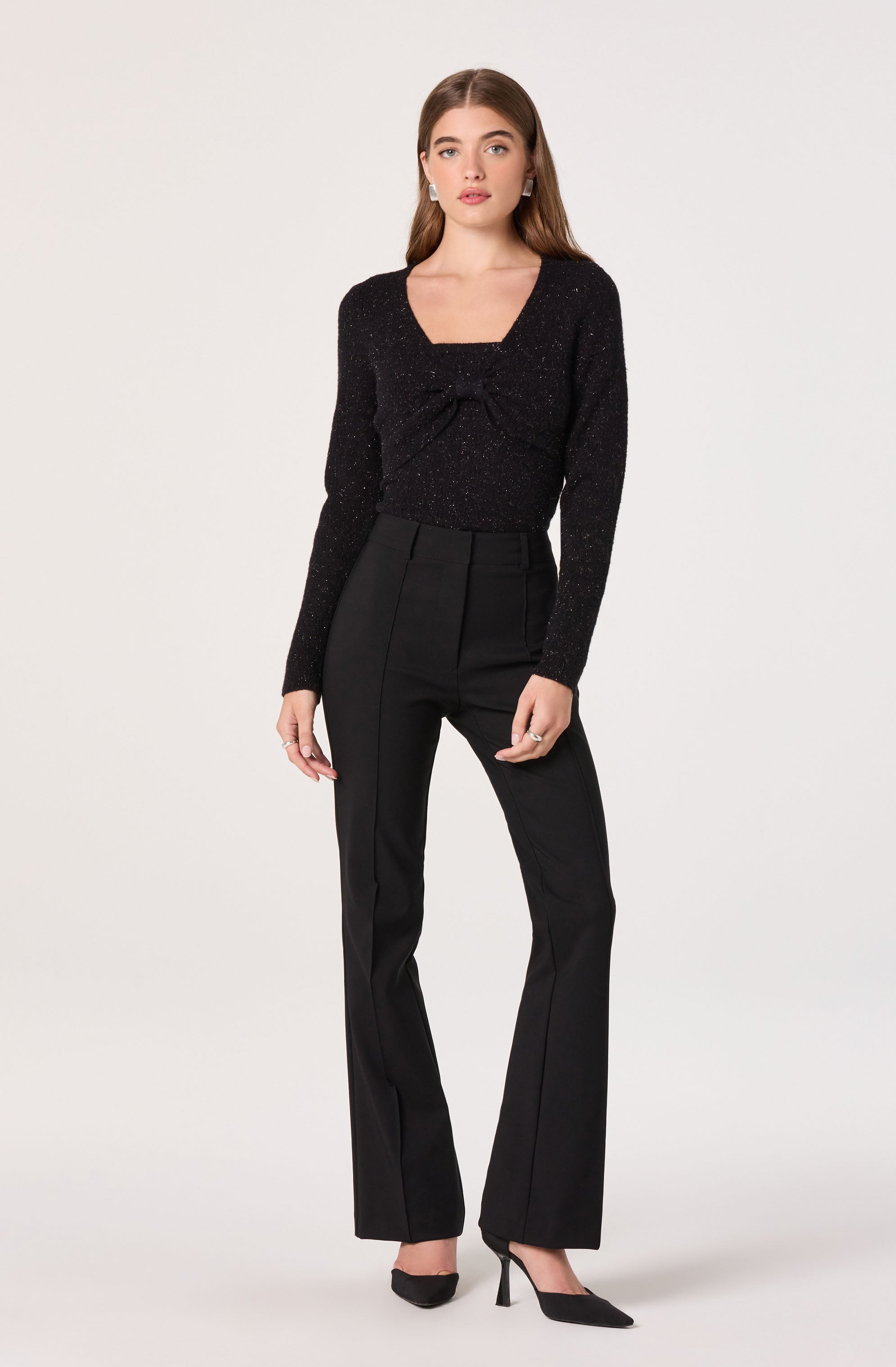 The woman is wearing a black, sparkly long-sleeve top with a square neckline and a decorative bow detail in the front. She is also dressed in high-waisted black trousers with a straight-leg cut, and black high-heeled shoes.