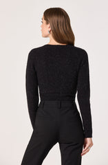 A woman is wearing a black, long-sleeved, sparkly sweater with a round neckline, paired with high-waisted black tailored pants that have belt loops and back welt pockets.