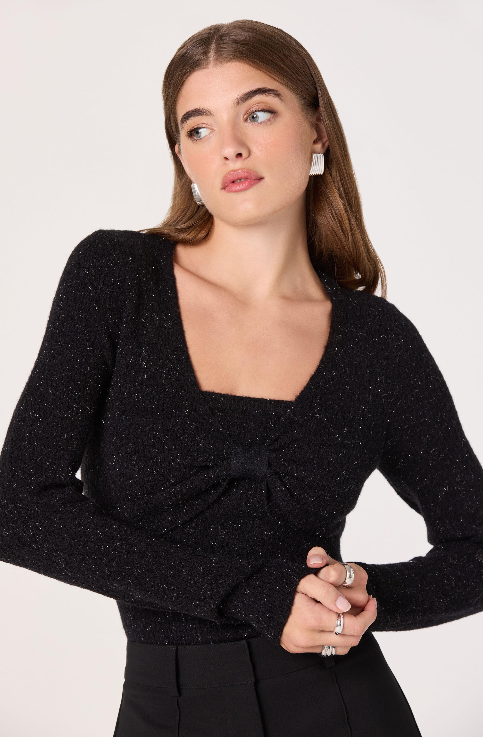 A woman is wearing a black long-sleeve top with a square neckline and a gathered detail at the bust. She is also wearing black high-waisted pants. Her accessories include large white rectangular earrings and multiple rings on her fingers.