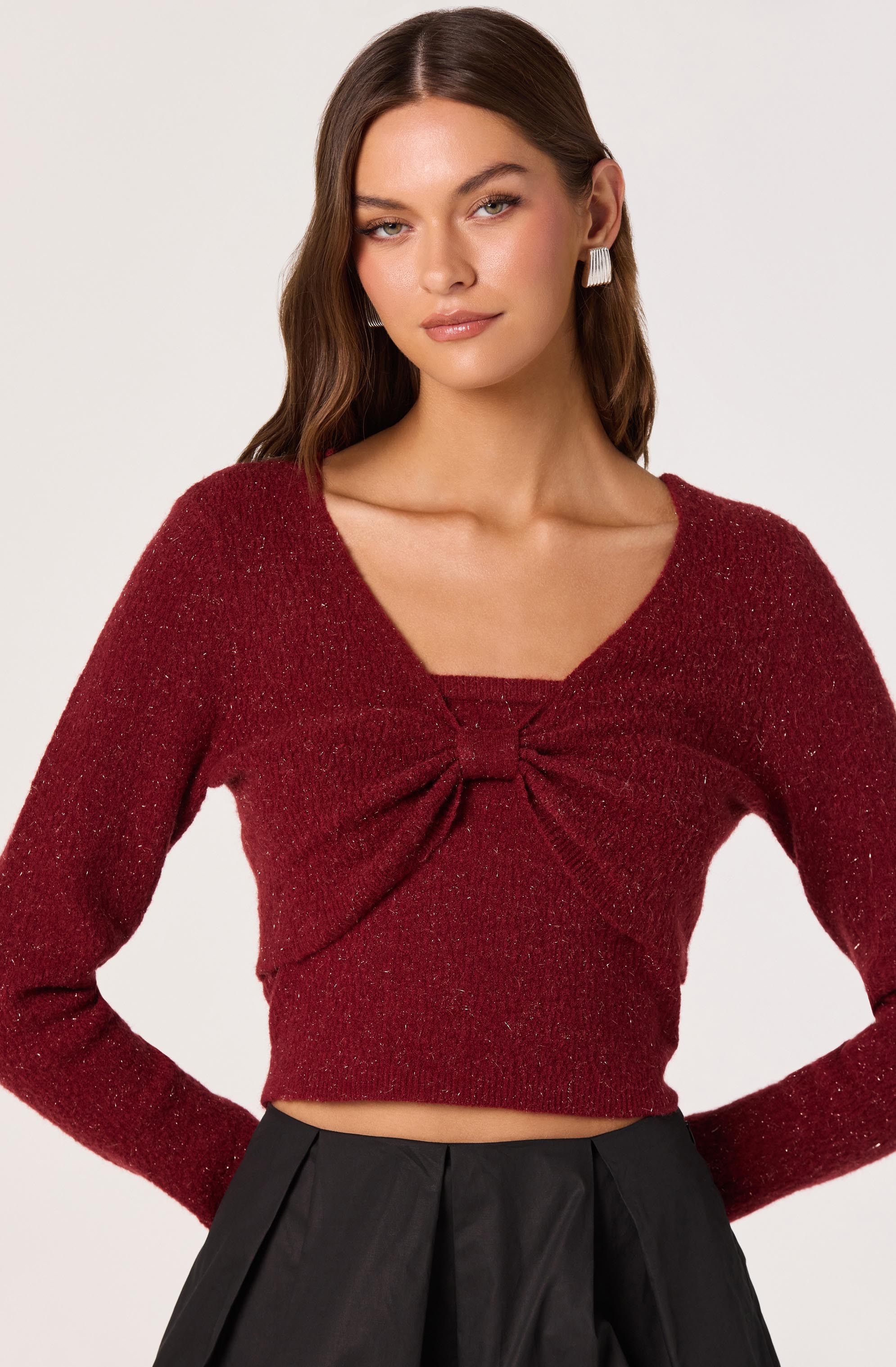 A woman is wearing a long-sleeve, deep red, textured sweater with a bow detail at the center of the chest. The sweater has a wide, rounded neckline and is cropped, ending just above the waist. She pairs it with high-waisted black pants that have pleats in the front. She is accessorized with large, rectangular white earrings.