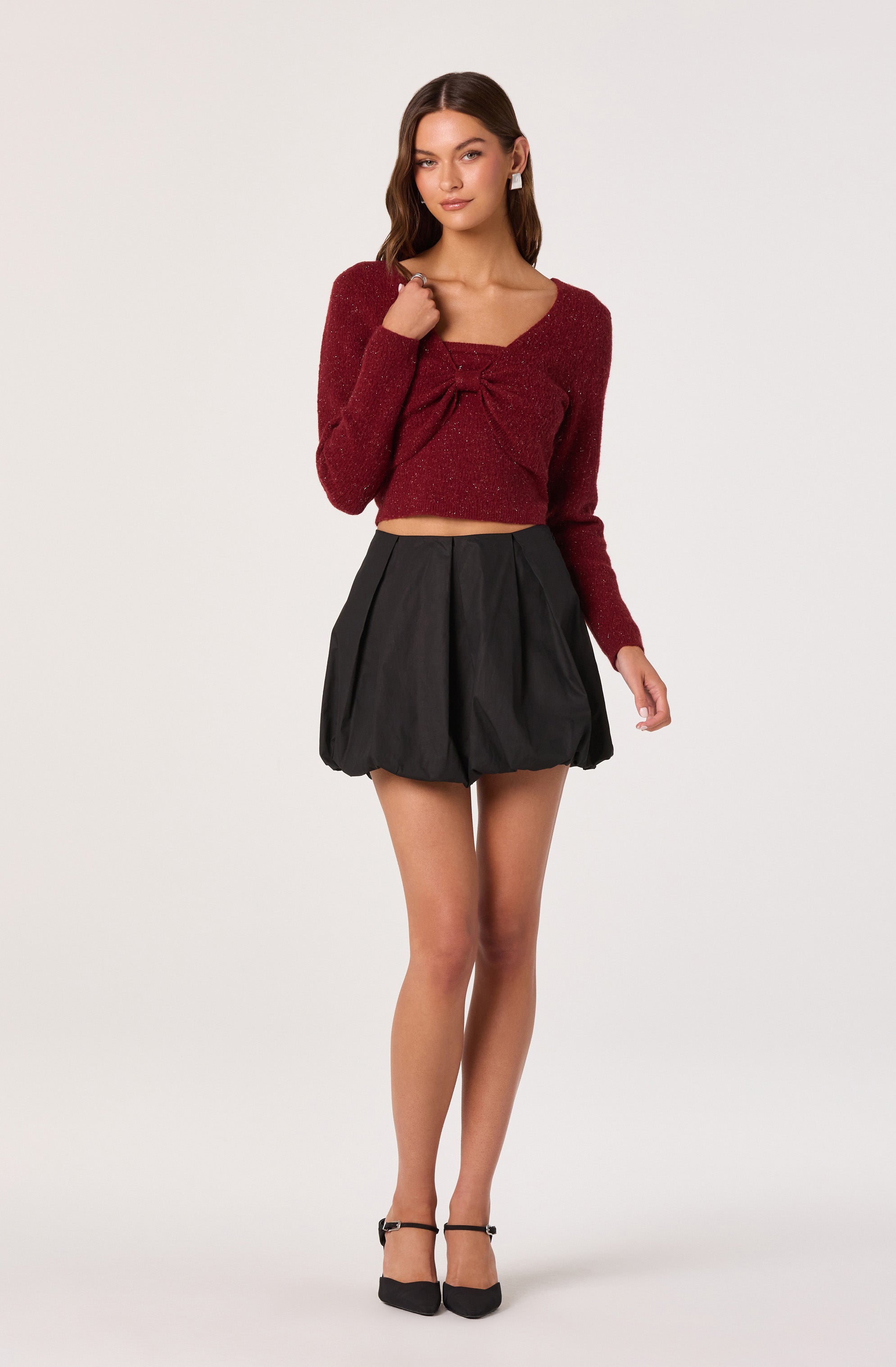 A young woman is wearing a long-sleeved, sparkly, deep red crop top with a gathered detail at the center of the neckline. She pairs it with a high-waisted, loose-fitting black mini skirt that has a slightly puffy silhouette. She completes the look with black ankle-strap heels.
