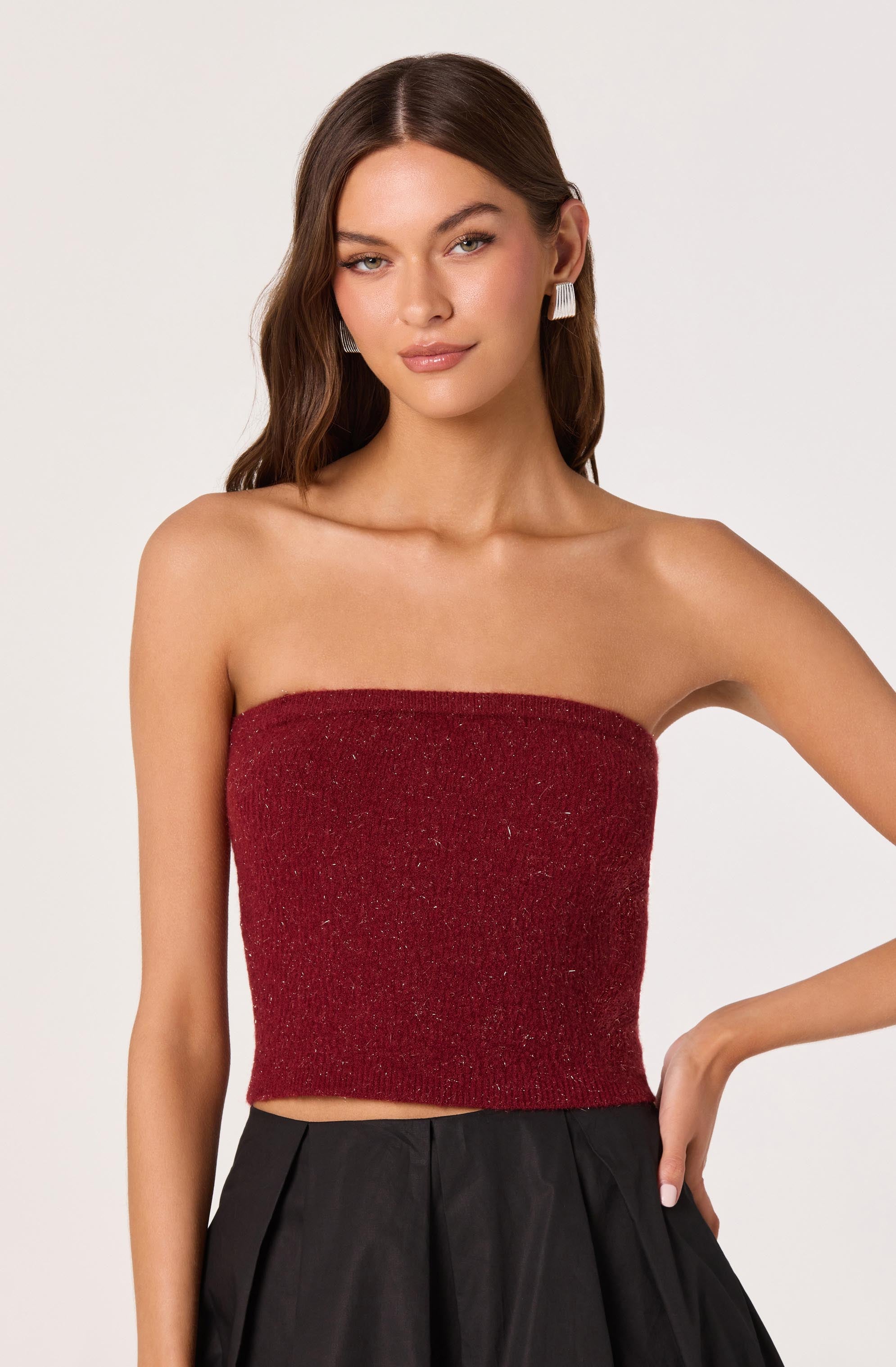 A woman is wearing a strapless, red, sparkly top that fits snugly around her torso. She has paired it with high-waisted black pants that feature pleats at the front. She is also wearing large, rectangular earrings.