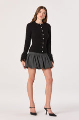 A woman is wearing a black, long-sleeved, button-up cardigan with a textured, fuzzy fabric and feathered cuffs. She is also wearing a high-waisted, dark gray, voluminous mini skirt with a gathered, bubble style. She has on black high-heeled shoes with ankle straps.