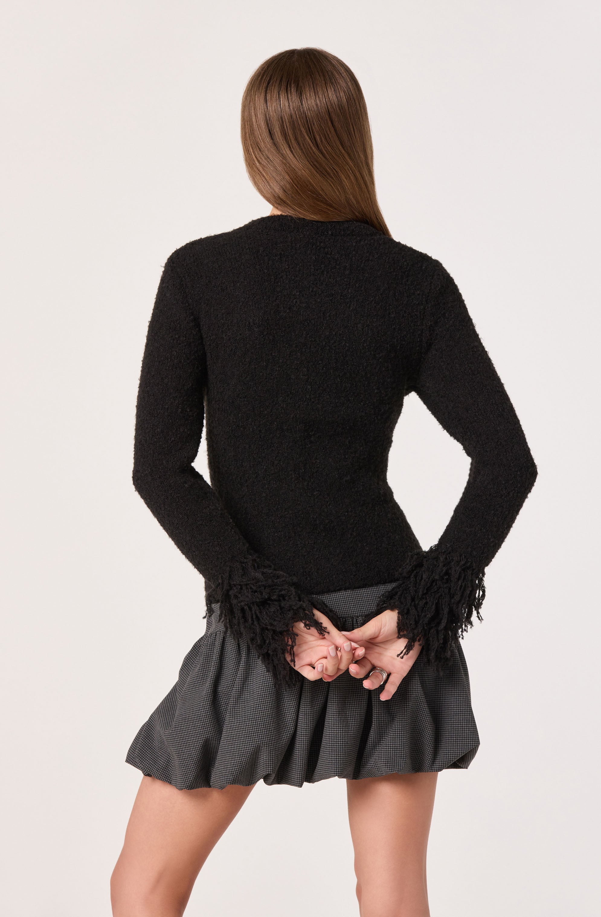 A person is wearing a black, textured sweater with long sleeves that have fringed details at the cuffs. They are also wearing a high-waisted, pleated gray skirt with a subtle checkered pattern. The person's hair is long and straight.