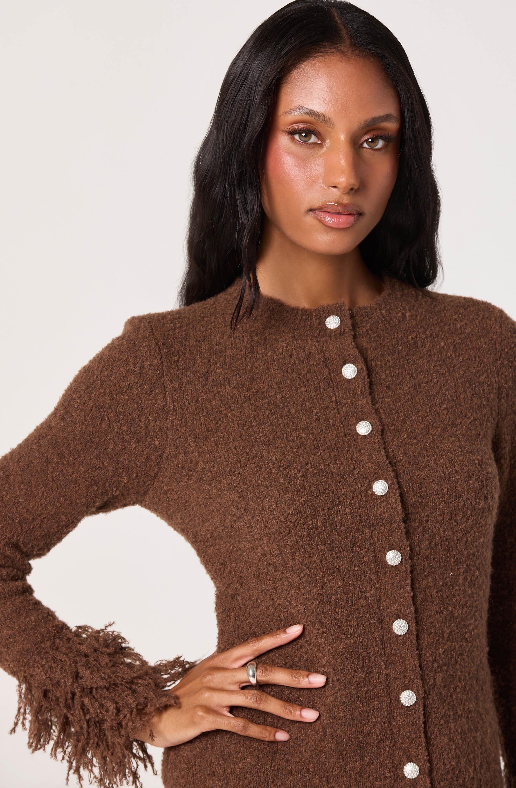 A woman is wearing a brown, long-sleeved, textured knit sweater with a round neckline. The sweater features a row of white buttons down the front and has fringed details on the cuffs.