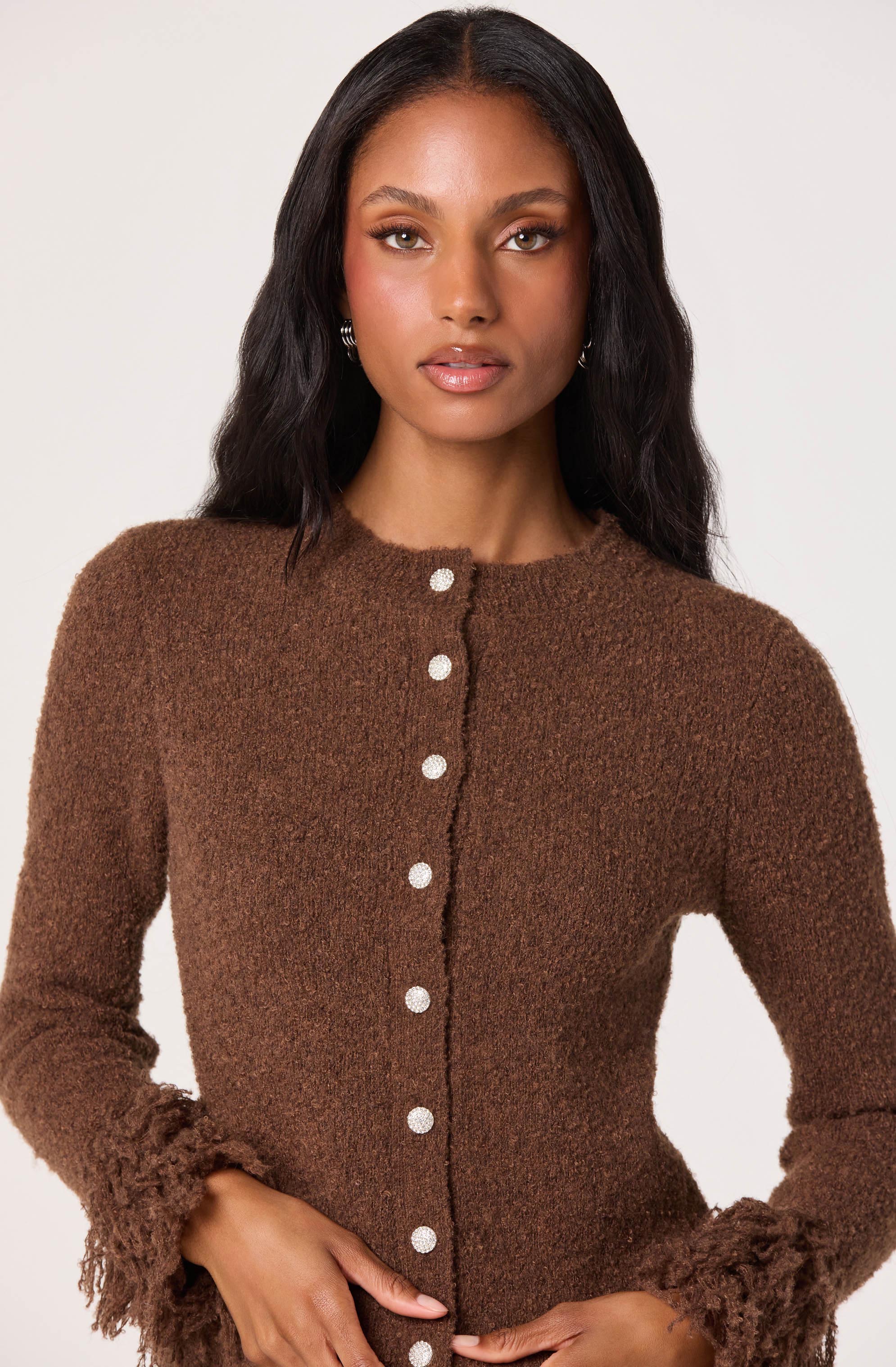 A woman is wearing a brown, textured cardigan with a fuzzy, slightly ruffled trim at the cuffs and hem. The cardigan features a row of white buttons down the front.