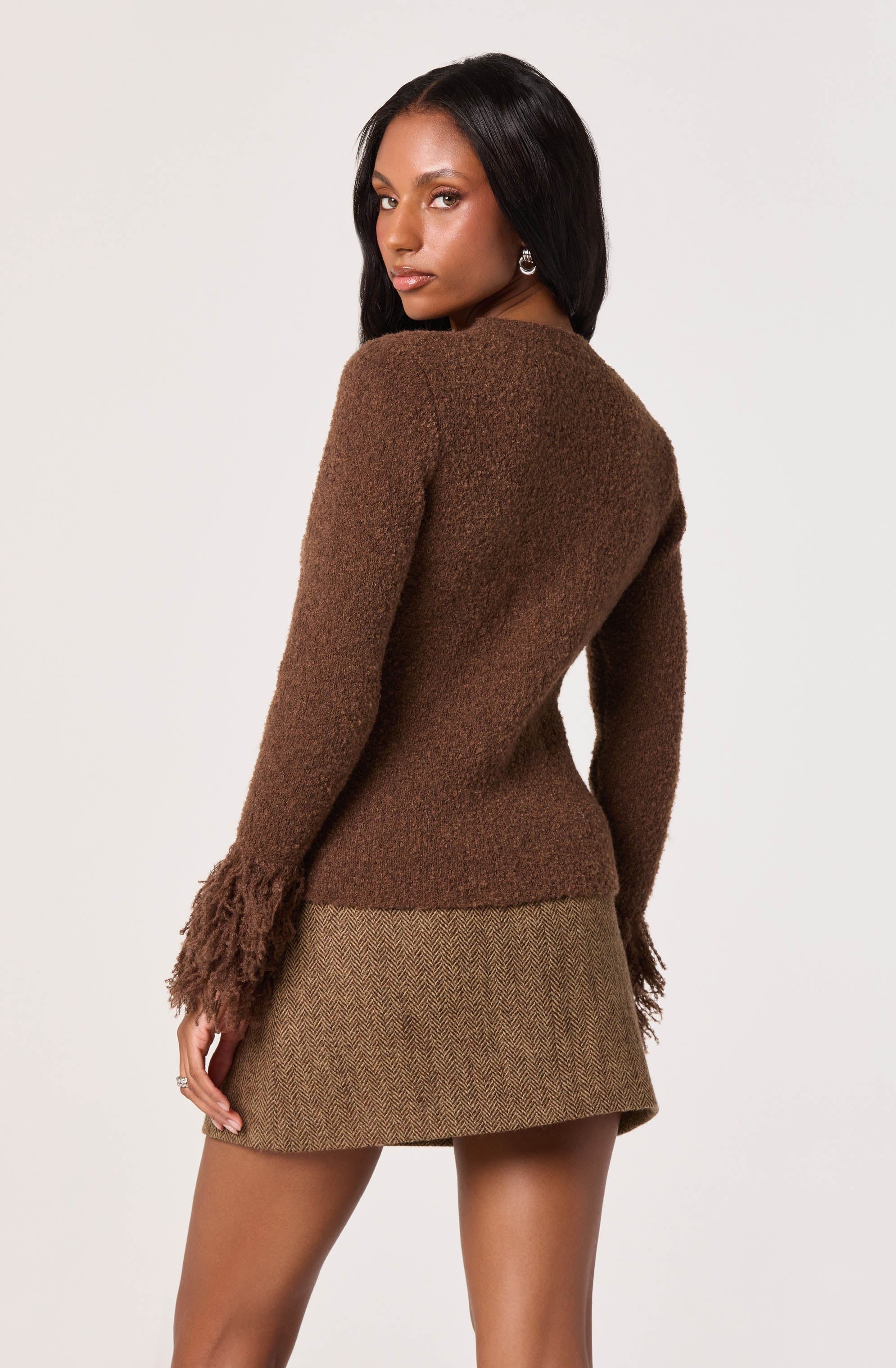 A woman is wearing a long-sleeved, brown, fuzzy sweater with textured, fringed cuffs at the wrists. She is also wearing a high-waisted, short skirt with a subtle herringbone pattern in muted earth tones.