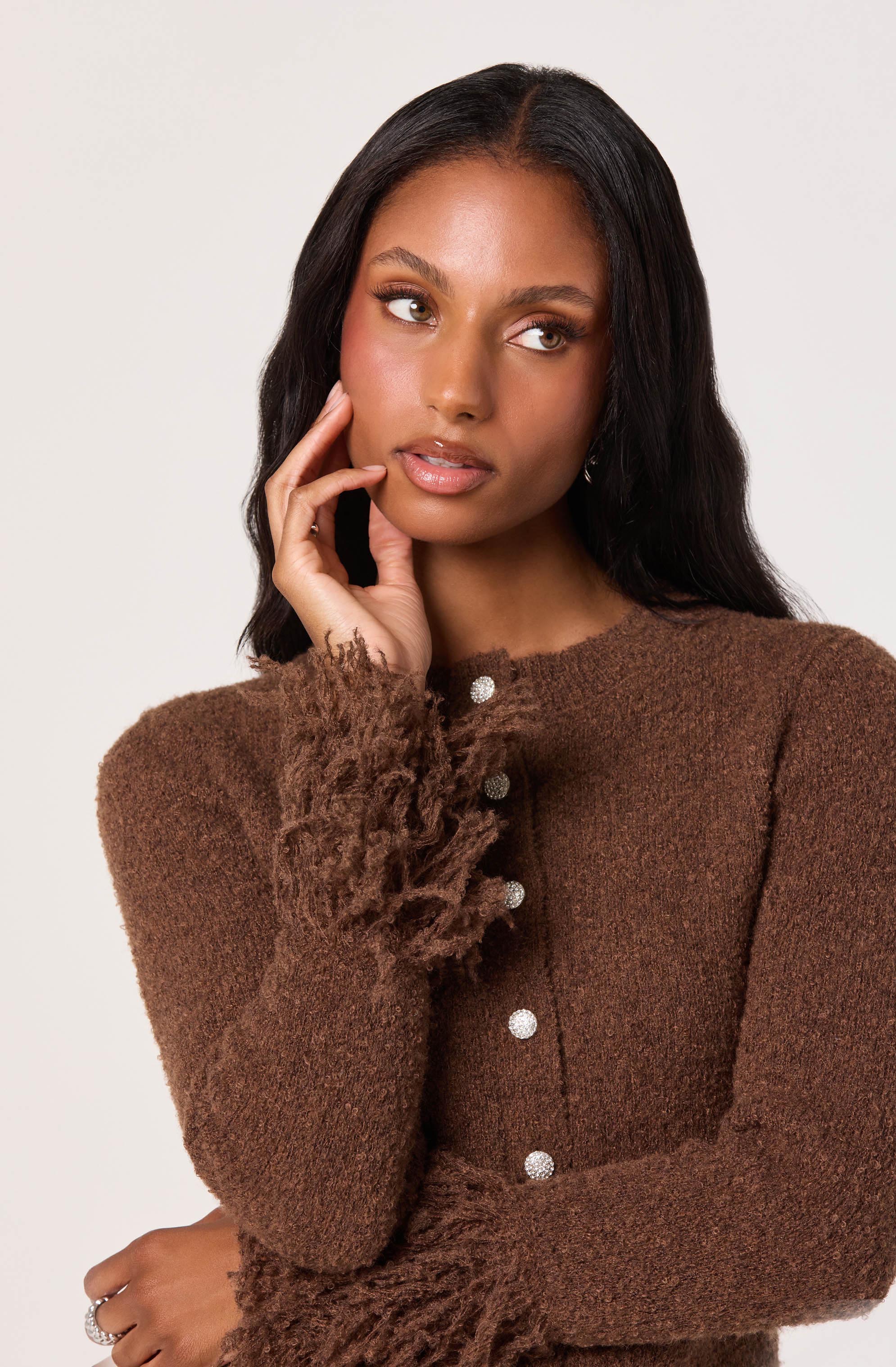 A woman is wearing a brown, textured sweater with a fuzzy trim on the cuffs and down the front, which features a row of shiny, round buttons.