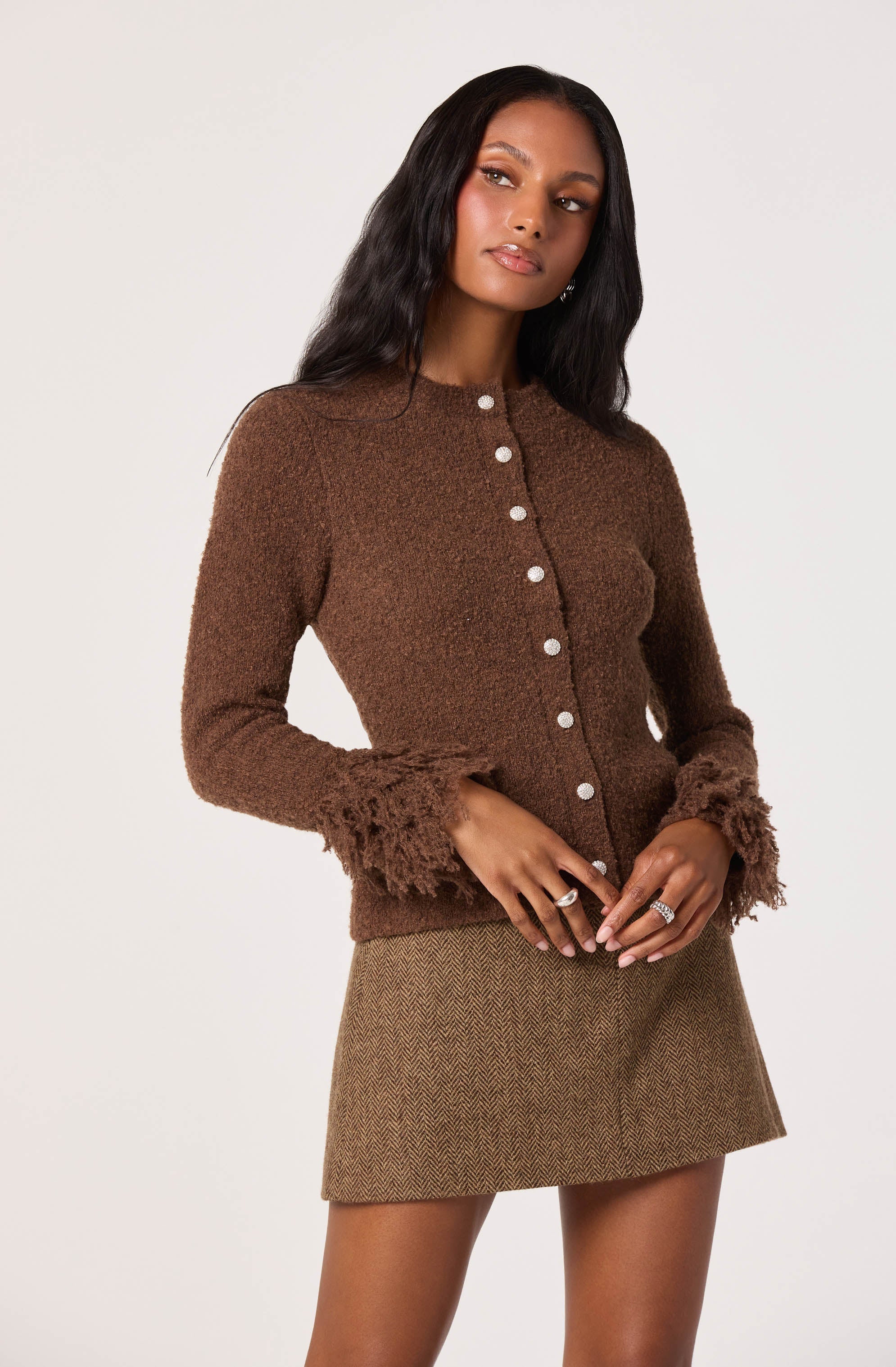 A woman is wearing a fitted brown cardigan with textured, fuzzy cuffs and buttons down the front. She is also wearing a short, brown checkered skirt.