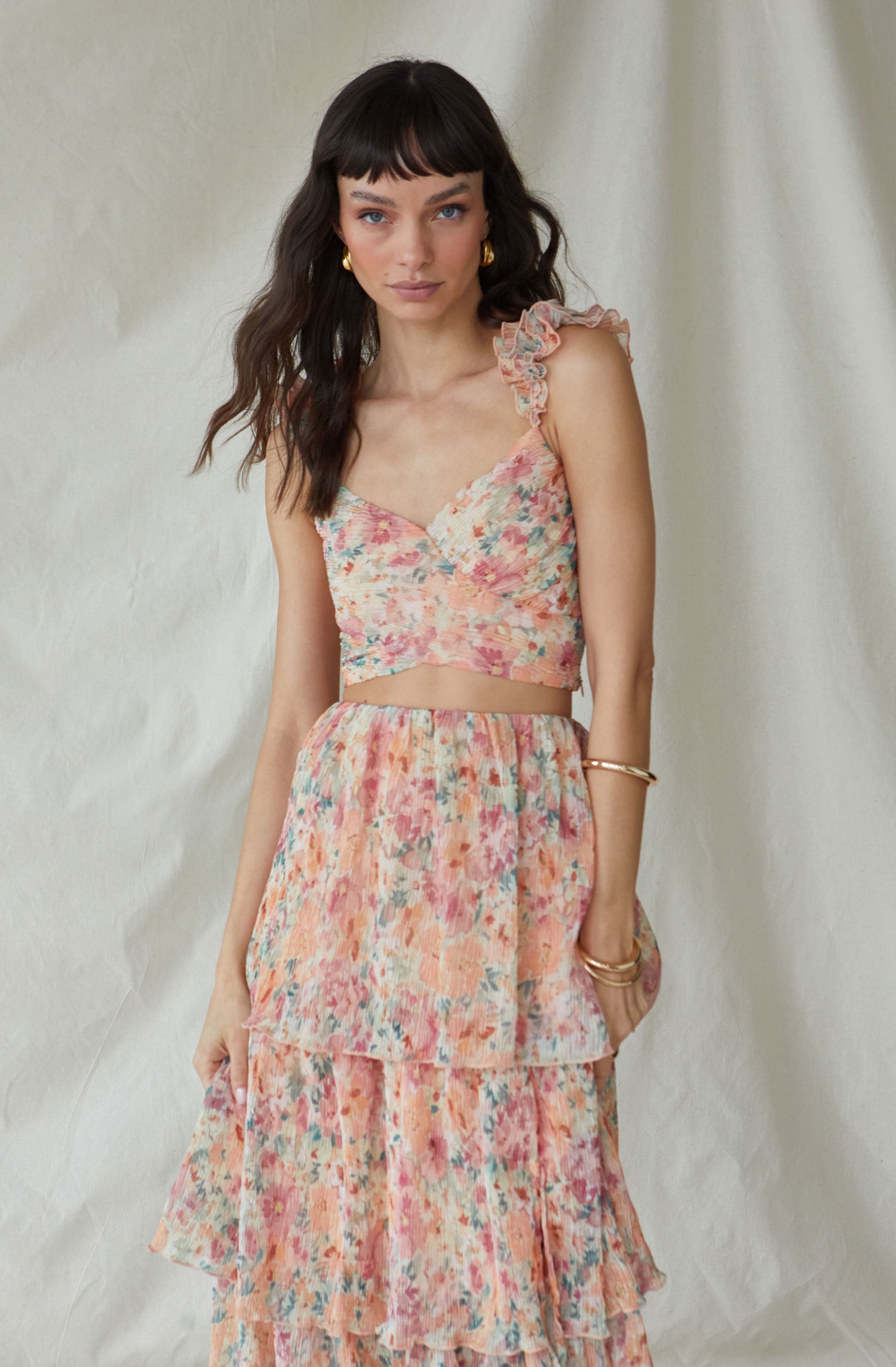 The woman is wearing a sleeveless crop top with ruffled shoulder straps, featuring a colorful, abstract floral print in shades of pink, peach, and light blue. She is also dressed in a matching layered skirt with the same vibrant, watercolor-like floral pattern.