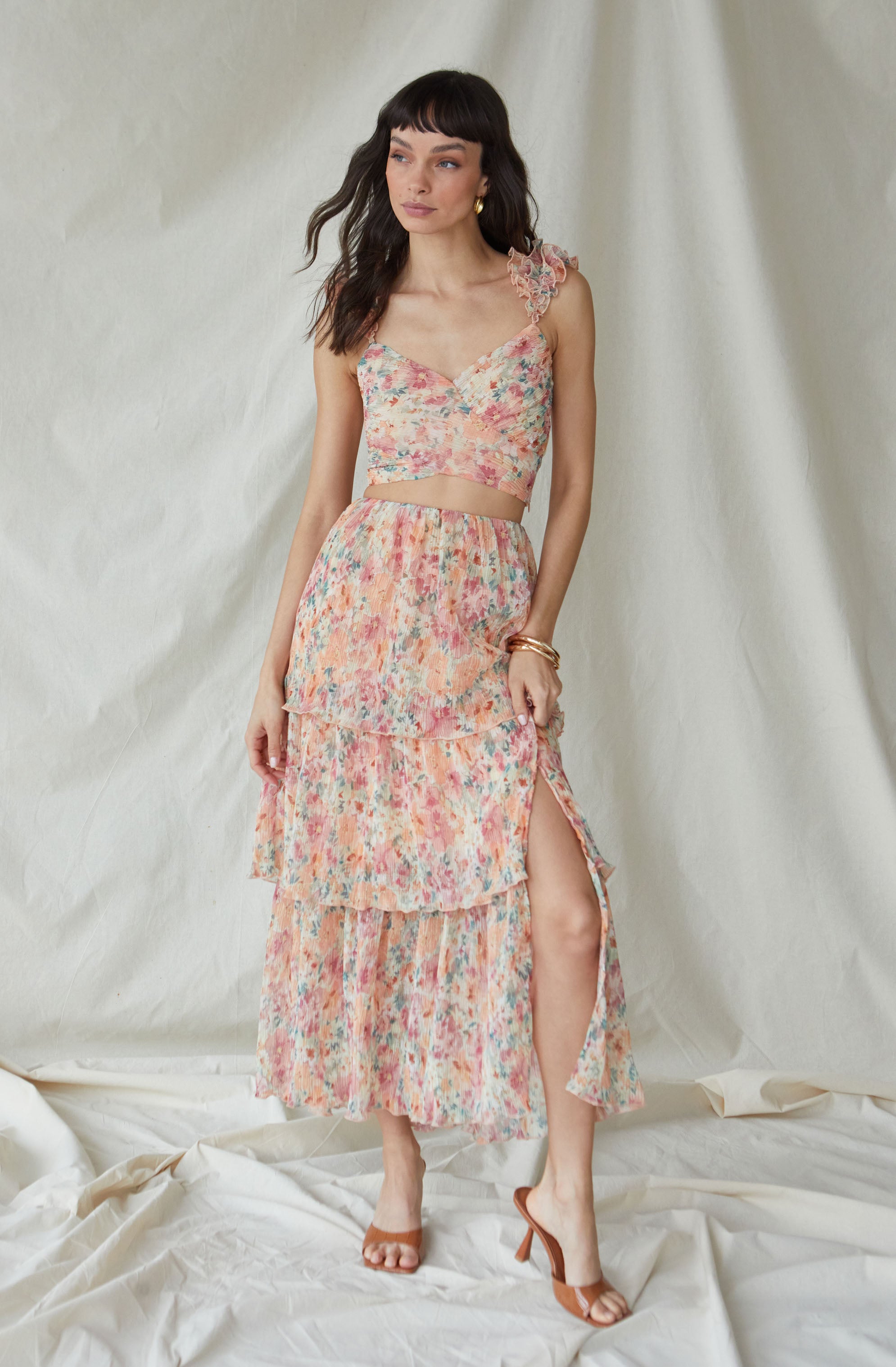 The outfit features a sleeveless crop top with a floral pattern and ruffled shoulder straps. The matching skirt has a layered, tiered design with a slit on one side, revealing a high slit up to the thigh. The fabric of both pieces has a lightweight, flowy texture with a multicolored, pastel floral print. The model accessorizes with gold earrings and bracelets, and wears strappy high-heeled sandals.