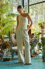 A woman is wearing a sleeveless, one-shoulder cream-colored top with button details down the front, paired with high-waisted, wide-leg cream-colored trousers. She accessorizes with a chunky gold chain necklace and light-colored open-toe shoes. The setting is a bright, plant-filled room with large windows, lush greenery, and various potted plants and flowers surrounding her.