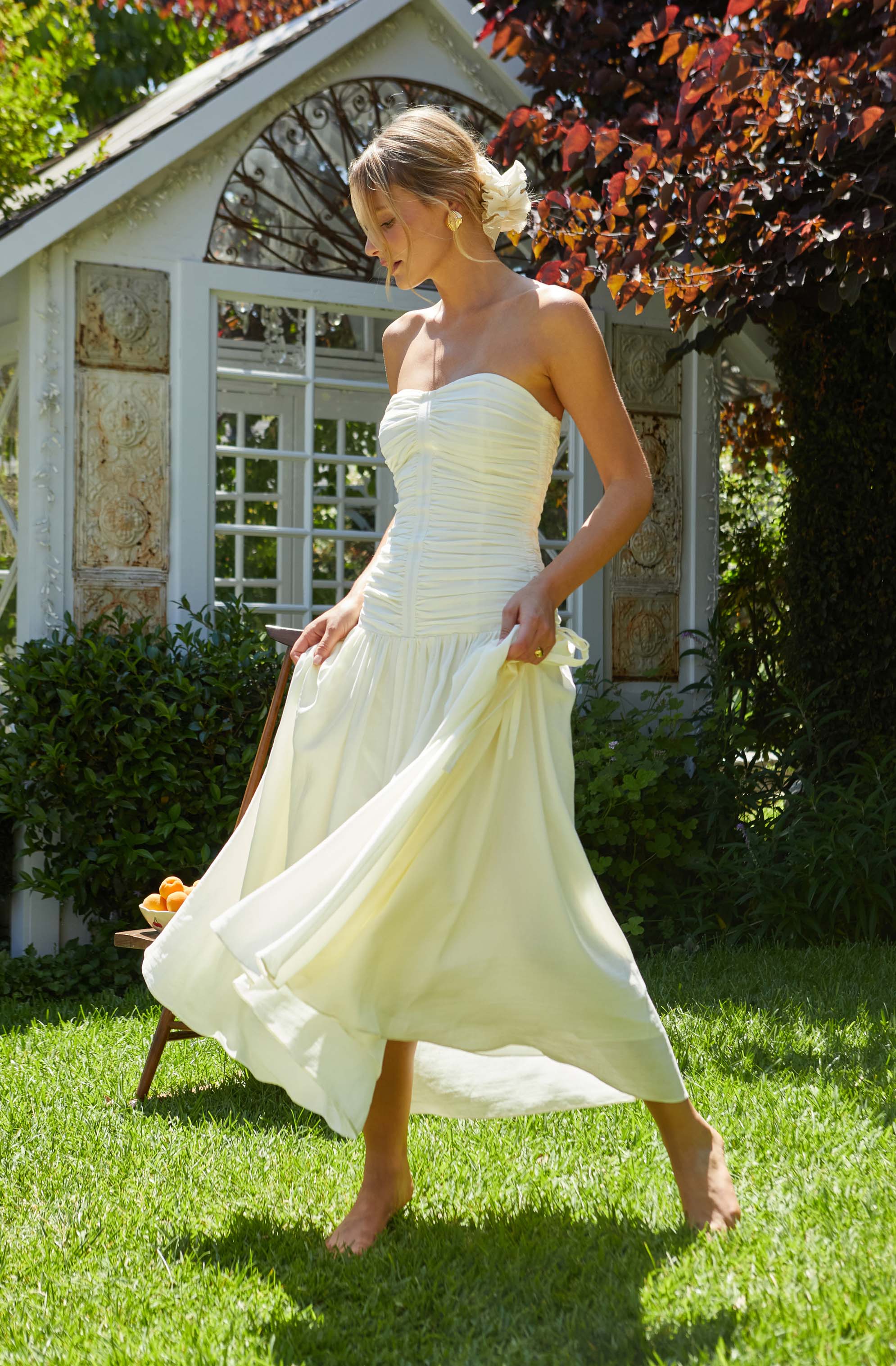 A woman is wearing a strapless, cream-colored dress with a fitted, ruched bodice and a flowing, full skirt. The dress is elegant and appears suitable for a formal occasion or outdoor event. She has a white flower in her hair and stands barefoot on a grassy lawn with a garden and a decorative white structure in the background.