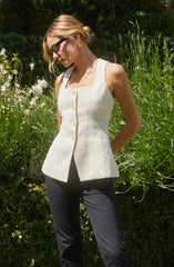 A woman is wearing a sleeveless, off-white, textured button-up top with large buttons down the front. The top has a square neckline and a fitted silhouette. She pairs it with dark-colored pants. She accessorizes with large black sunglasses, gold earrings, and a gold bracelet.