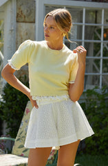 A woman is wearing a light yellow, short-sleeved knit top with a round neckline and lace trim on the sleeves and bottom hem. She is also wearing a white, high-waisted, A-line skirt with a perforated eyelet pattern.