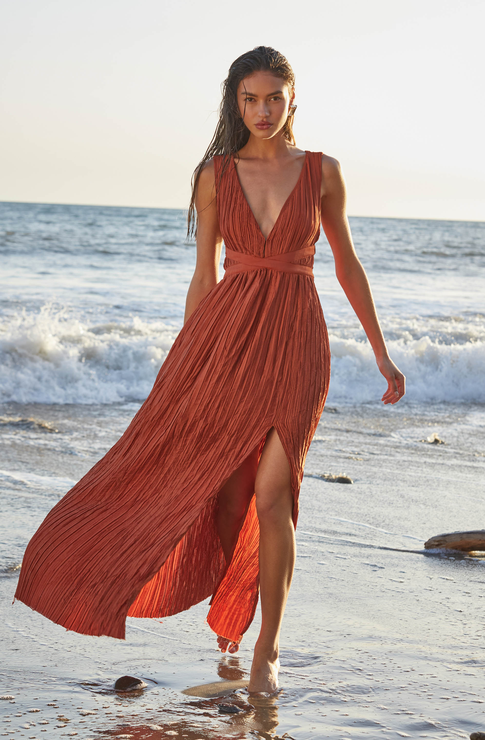 A woman is wearing a sleeveless, deep V-neck, rust-colored pleated maxi dress with a fitted waist and a high slit on the left side, walking barefoot along the shoreline with waves in the background.