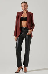 A woman is wearing a burgundy blazer with a relaxed fit and slightly padded shoulders over a black crop top. She has on high-waisted black leather pants and black high-heeled sandals.