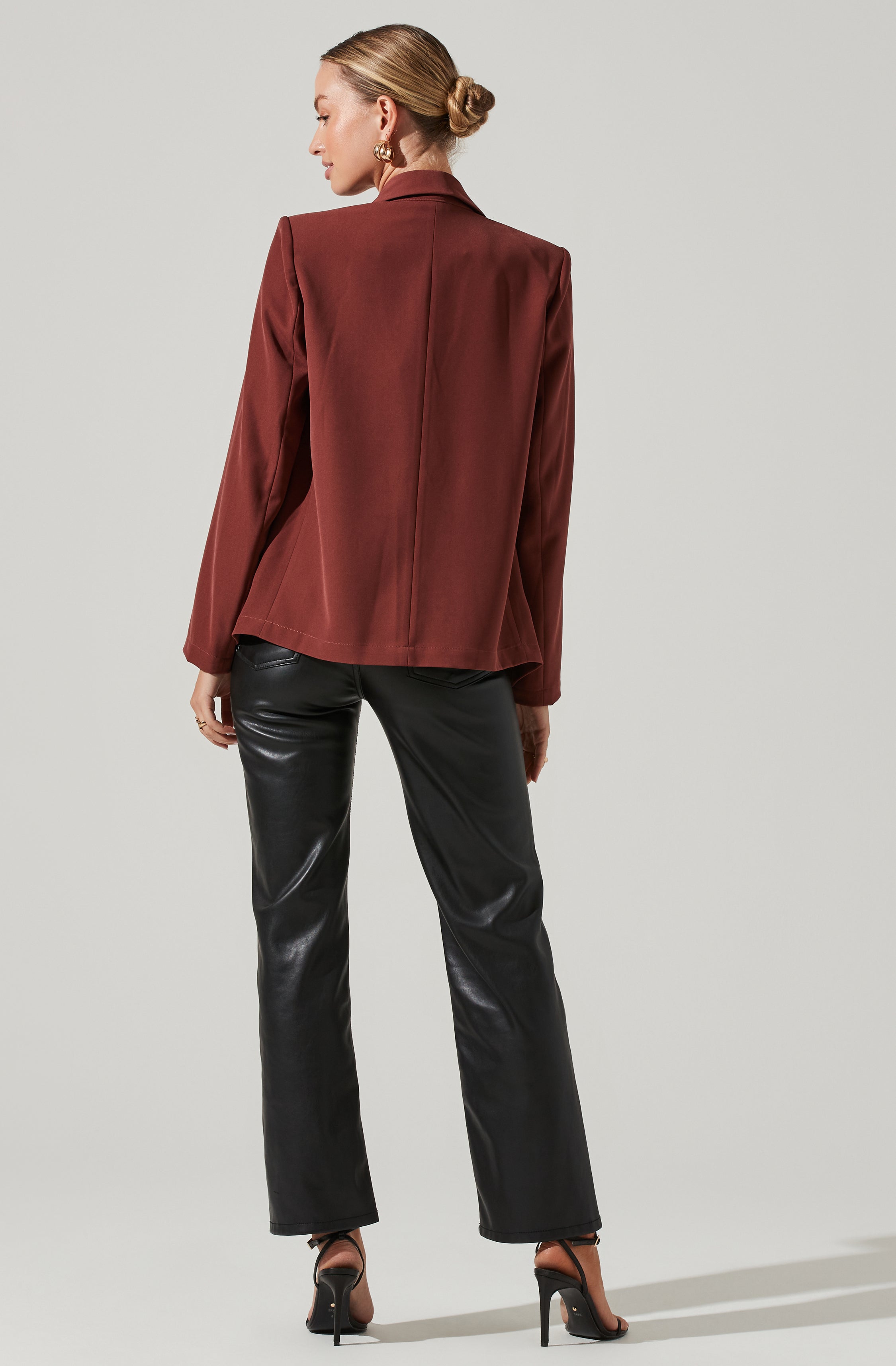 A woman is wearing a fitted, long-sleeved blazer in a deep reddish-brown color with a lapel collar, paired with black, shiny leather pants, and black high-heeled shoes.