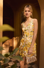 A young woman is wearing a sleeveless, V-neck dress adorned with large, embroidered floral patterns in shades of yellow, white, and green. She is holding a metallic gold handbag with gathered fabric detail and is standing in a warmly lit room with a checkered black and white floor, wooden doorway, and yellowish walls.