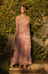 A woman is wearing a strapless, floor-length dress with a pastel floral pattern in shades of pink, peach, and green. The dress features a fitted bodice and a flowing skirt with ruffled detailing toward the bottom. She is holding a glittery clutch bag in her left hand.