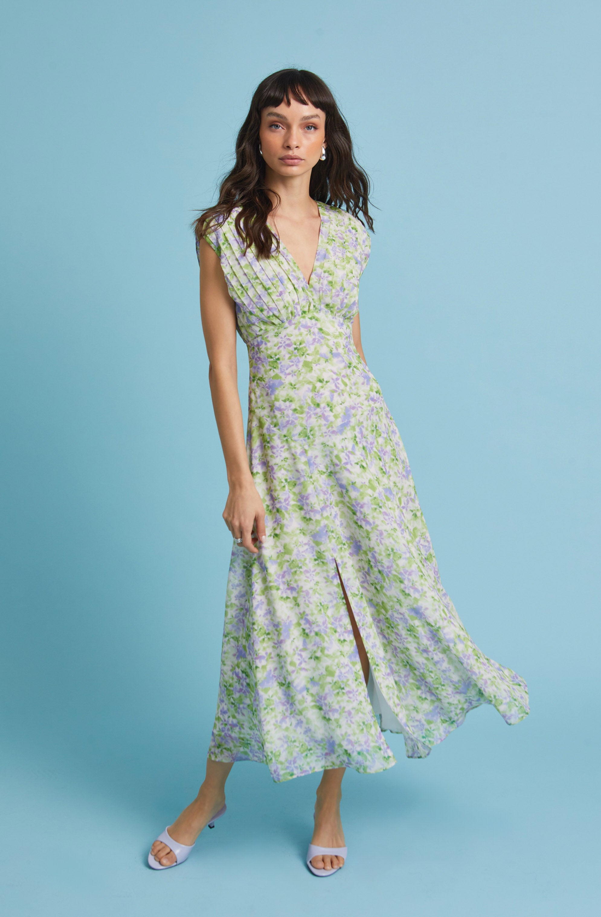 A woman is wearing a sleeveless, floor-length dress with a V-neckline. The dress features a floral pattern in shades of light purple, green, and white. It has a fitted waist and a front slit that reveals her legs. She is also wearing white open-toe heels.
