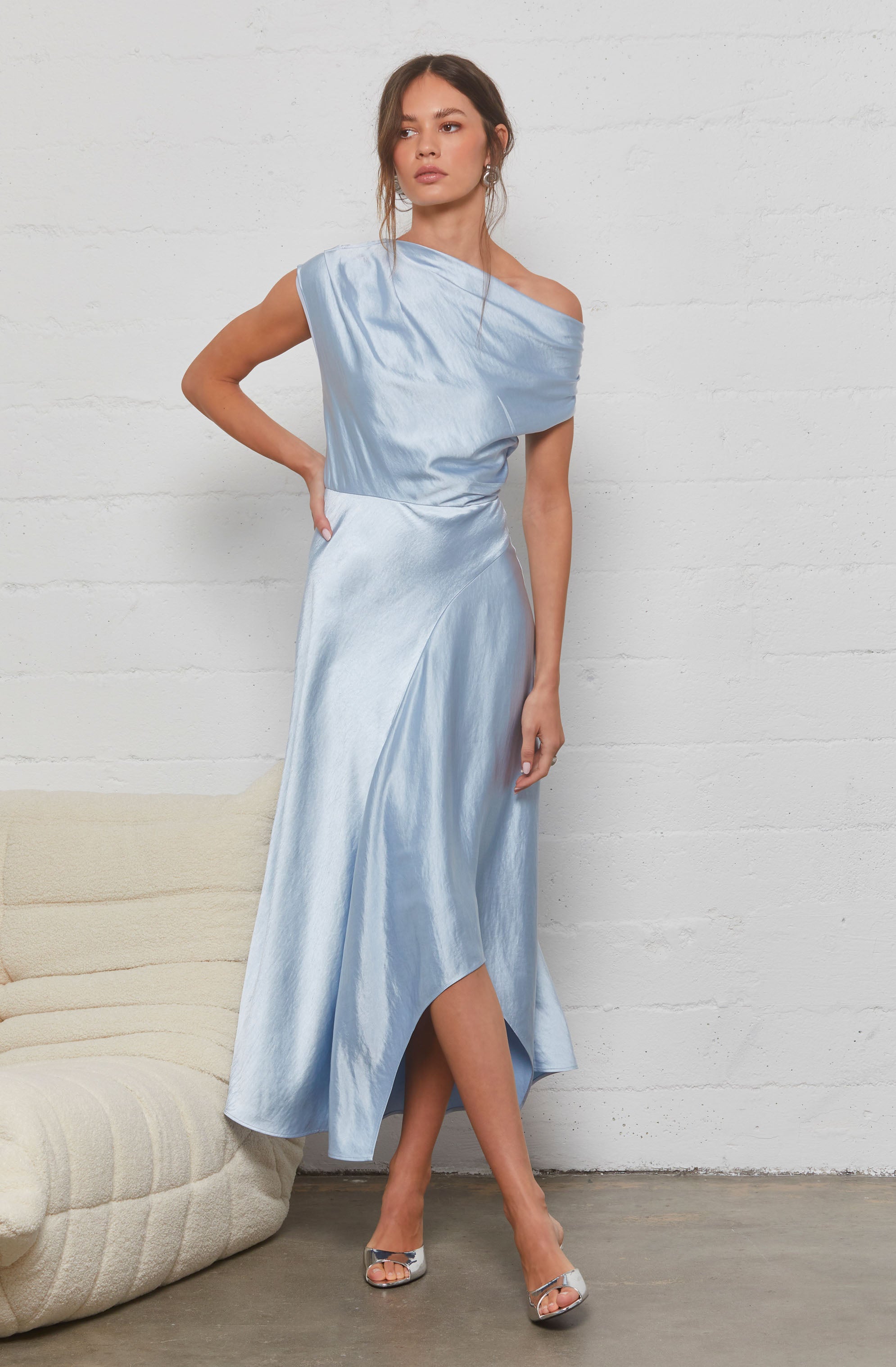 A woman is wearing a light blue, satin-like evening gown. The dress features an asymmetrical, off-the-shoulder neckline on one side and a sleeveless design on the other. The fabric has a smooth, shiny texture, and the gown has a fitted waist with a flowing, A-line skirt that reaches mid-calf with a slit on one side. She is also wearing silver high-heeled sandals.