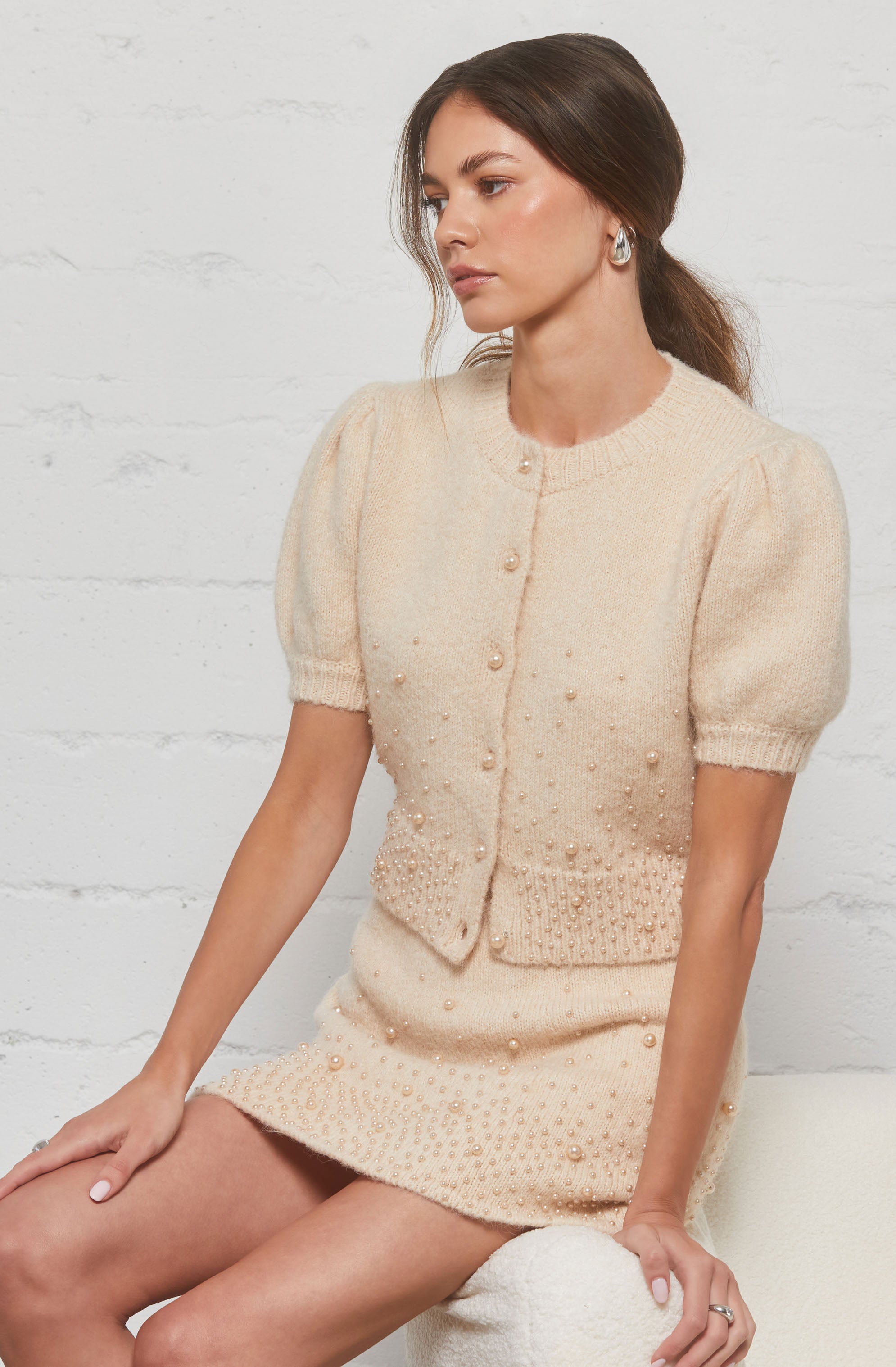 The woman is wearing a cream-colored, short-sleeved knit top with puffed sleeves and decorative pearl buttons down the front. The top has a textured design with small pearls scattered throughout and features a ribbed hem at the waist. She is also wearing a matching cream-colored knit skirt with similar pearl embellishments.