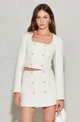 A woman is wearing a white, textured two-piece outfit consisting of a long-sleeved, cropped jacket with a rounded neckline and decorative buttons down the front, and a matching high-waisted mini skirt with similar buttons. She accessorizes with gold hoop earrings and holds a white handbag with a gold chain strap.