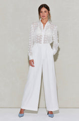 A woman is wearing a semi-sheer white blouse with long sleeves, featuring textured polka dot patterns and a classic collar. She pairs it with high-waisted, wide-leg white trousers that have pleats at the front. Her shoes are pointed-toe heels in a light blue color with a textured fabric.
