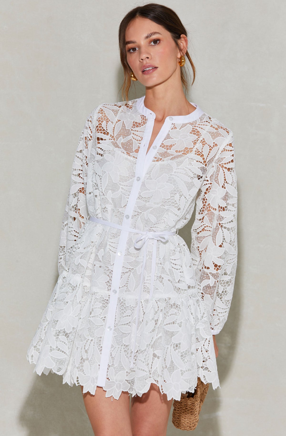A woman is wearing a white lace dress with floral patterns. The dress features long sleeves, a button-up front, and a waist tie. The lace has a delicate, intricate floral design, and the hemline is scalloped. She is accessorized with gold hoop earrings and holds a small woven bag.