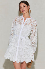 A woman is wearing a white lace dress with floral patterns. The dress features long sleeves, a button-up front, and a waist tie. The lace has a delicate, intricate floral design, and the hemline is scalloped. She is accessorized with gold hoop earrings and holds a small woven bag.