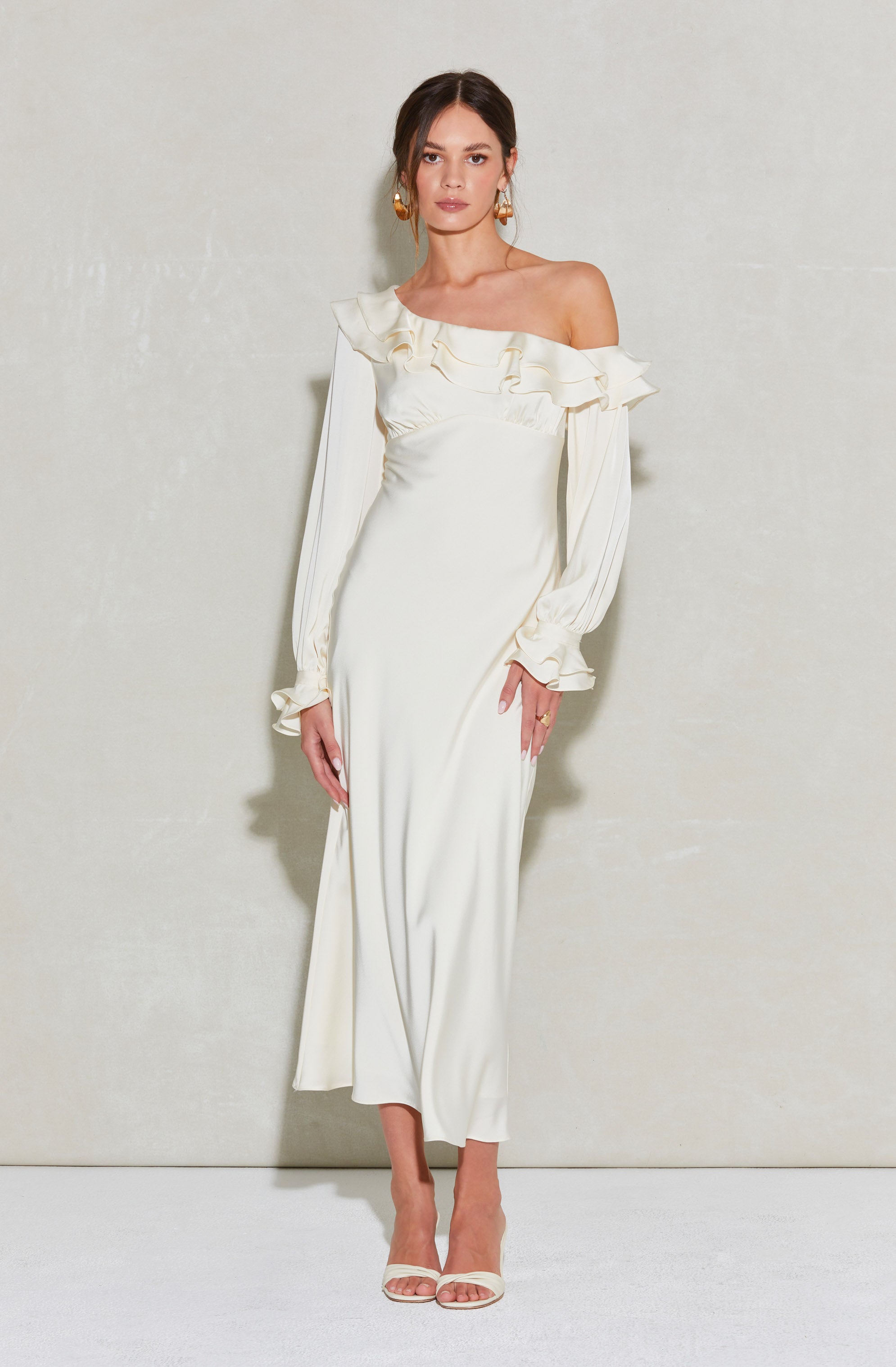 A woman is wearing an off-the-shoulder, cream-colored, full-length dress with ruffle detailing around the neckline and ruffled cuffs on the long sleeves. The dress is fitted and flows down to the floor. She is accessorized with large, gold hoop earrings and is wearing light-colored open-toe heels.