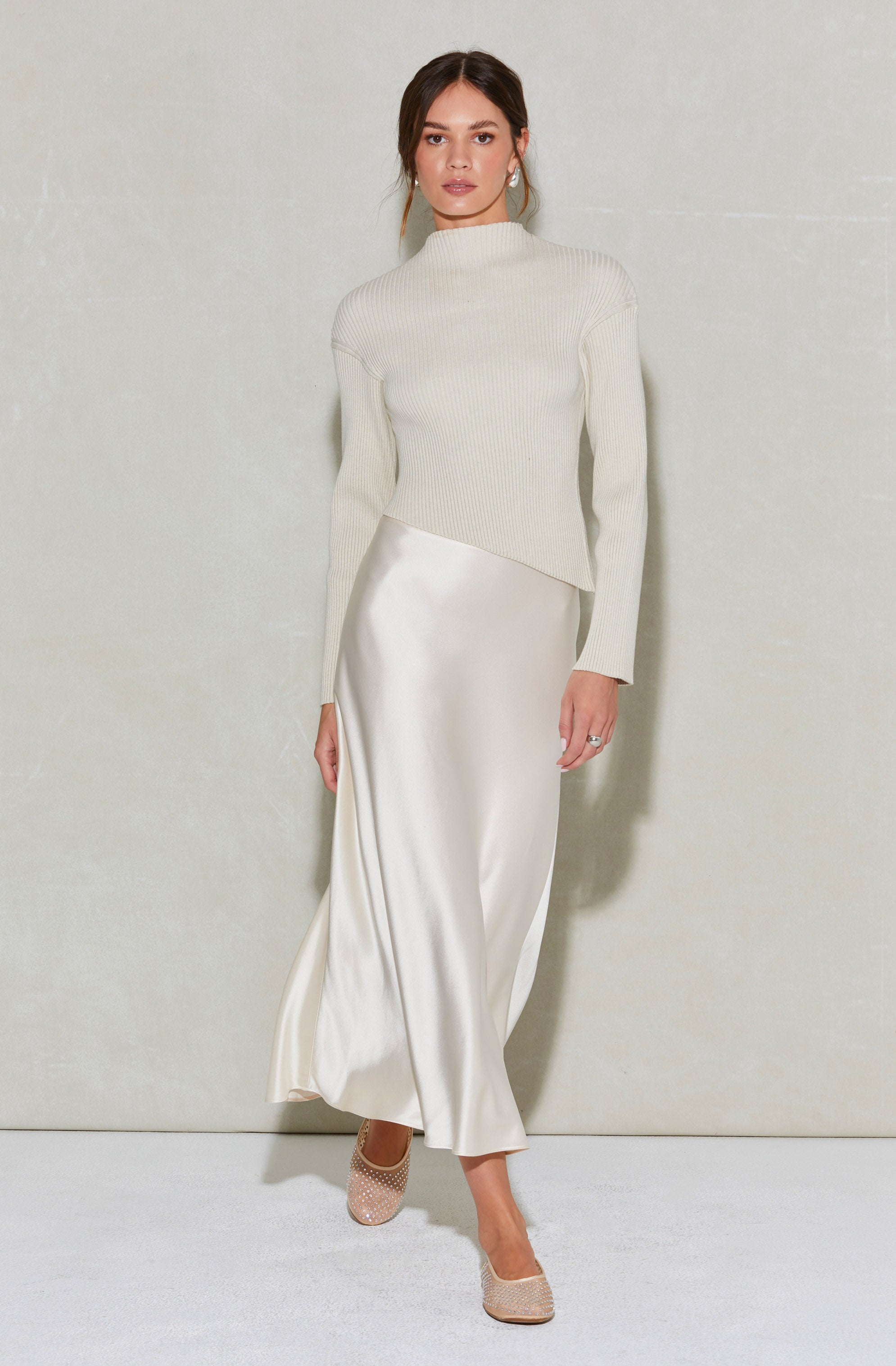 A woman is wearing a cream-colored, long-sleeved knit sweater with a high neckline. She pairs it with a long, flowing satin skirt that reaches her ankles. She completes the outfit with nude, peep-toe high heels.
