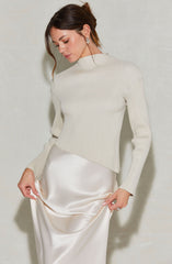 A woman is wearing a high-neck, long-sleeved ribbed sweater in a light, creamy color. She pairs it with a silky, satin-like skirt in an ivory shade that has a smooth, shiny texture.