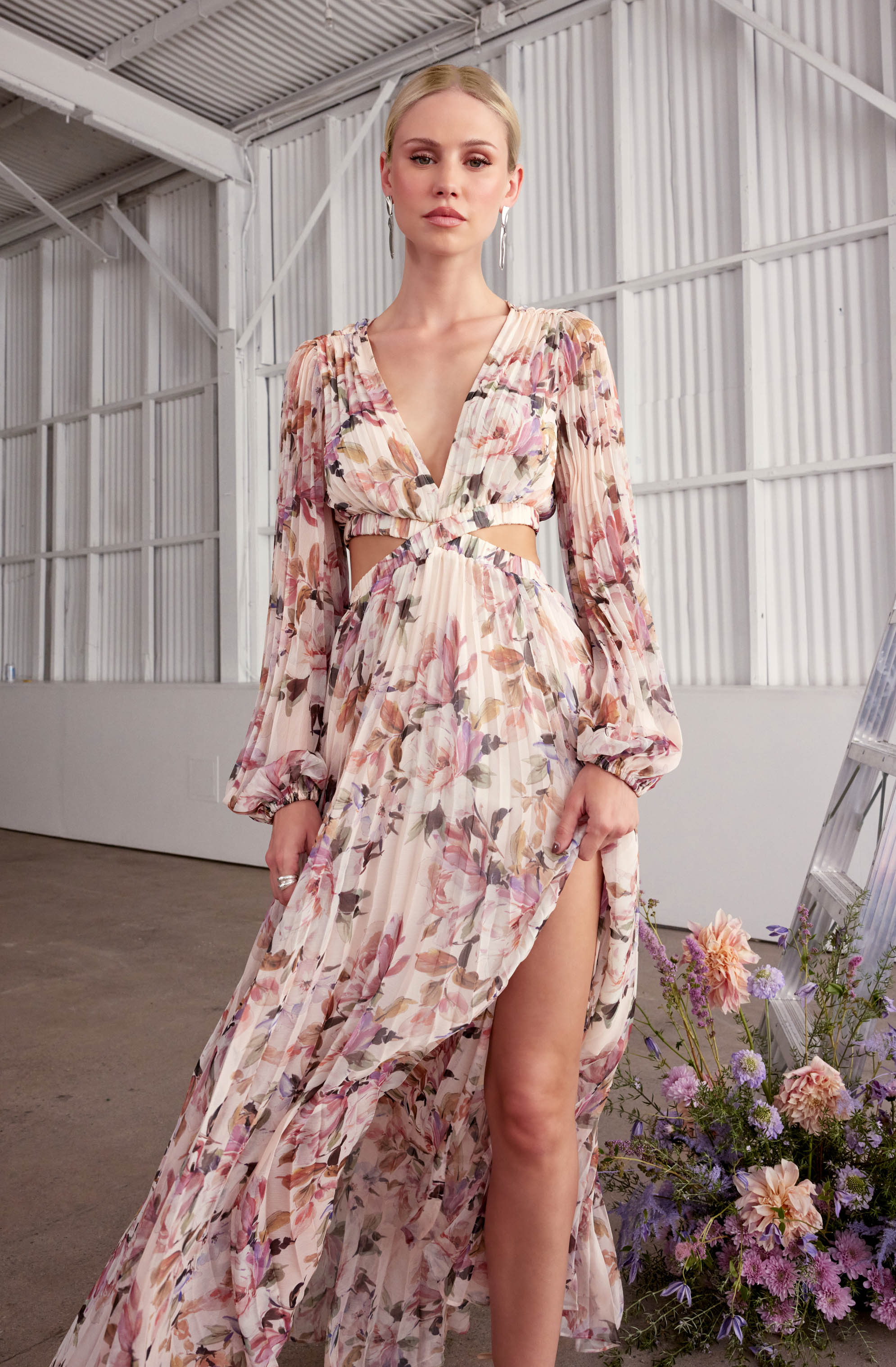 A woman is wearing a long, flowy dress with a pink and beige floral print. The dress features long, billowy sleeves with elastic cuffs, a deep V-neckline, and cut-out details at the waist. It also has a high slit revealing her leg. The fabric appears lightweight and sheer, with a gathered, ruffled skirt.