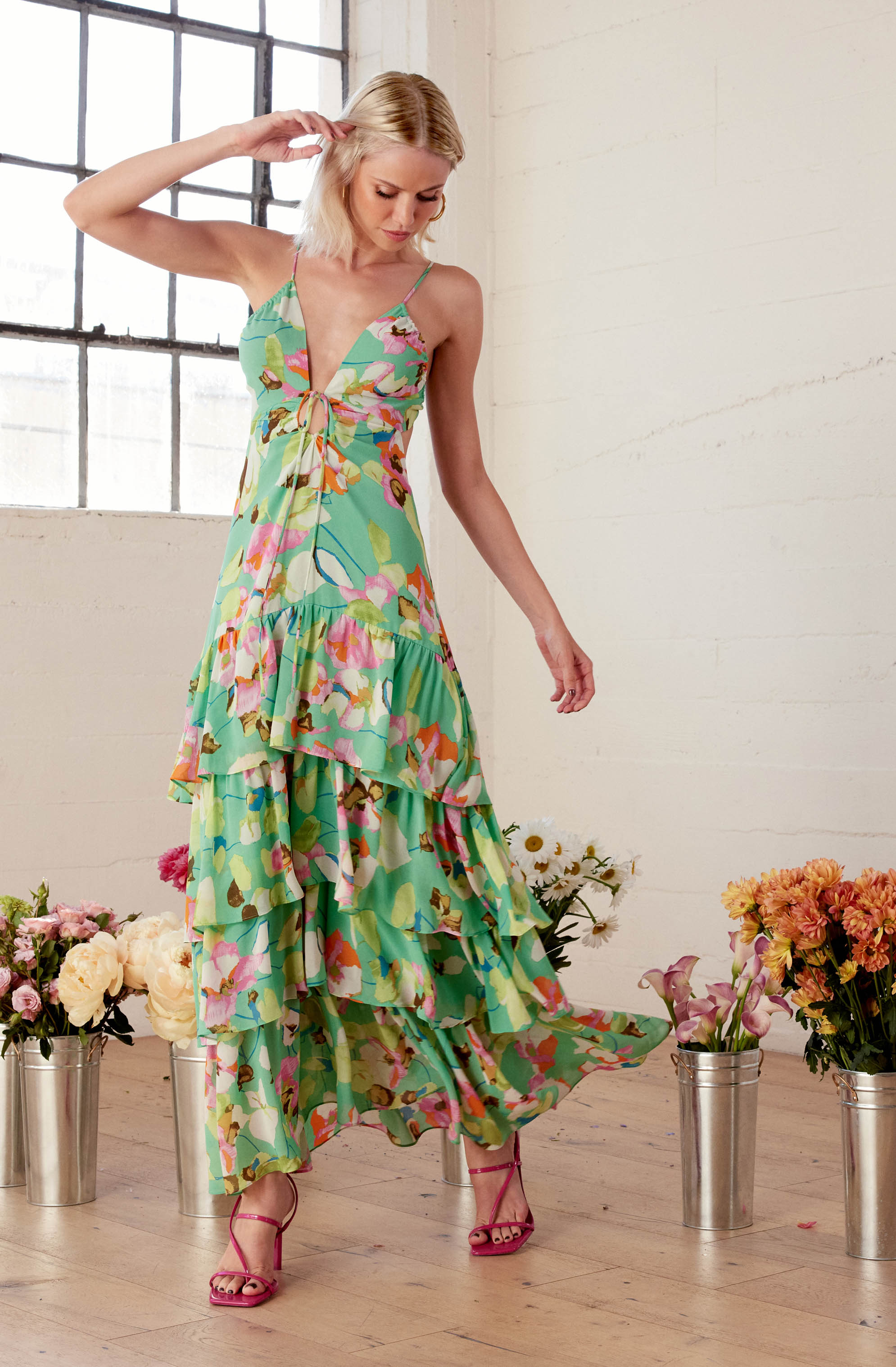 A woman is wearing a sleeveless, V-neck, tiered maxi dress with a colorful floral pattern in shades of pink, purple, orange, green, and blue. The dress features thin spaghetti straps and a tie detail at the front. She is also wearing high-heeled sandals in a bright pink color.