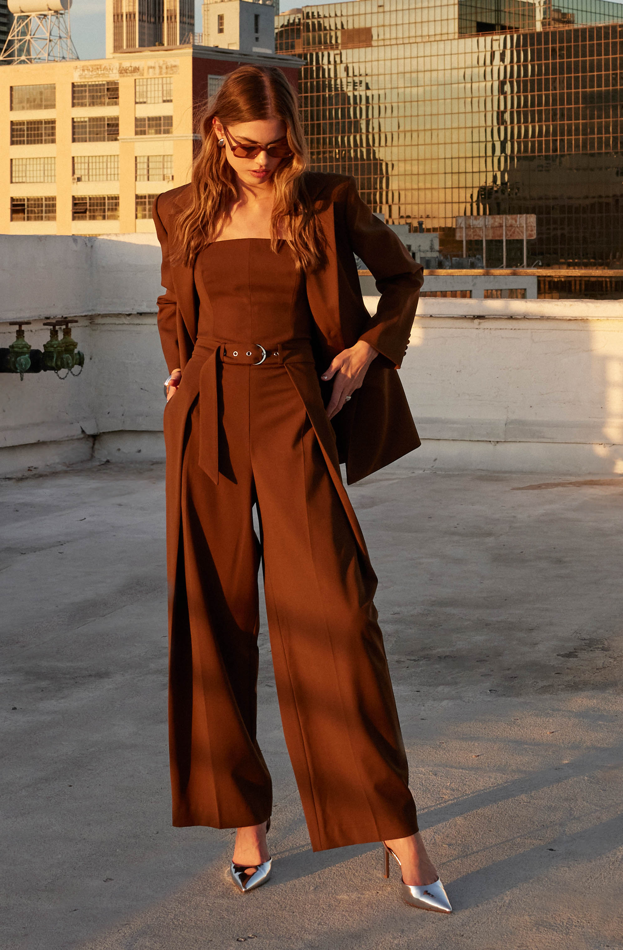 A woman is wearing a matching brown blazer and high-waisted, wide-leg trousers. The blazer is tailored and fitted at the waist, while the trousers are loose and flowing. She pairs the outfit with pointed metallic silver high heels. The setting appears to be a rooftop with city buildings in the background.