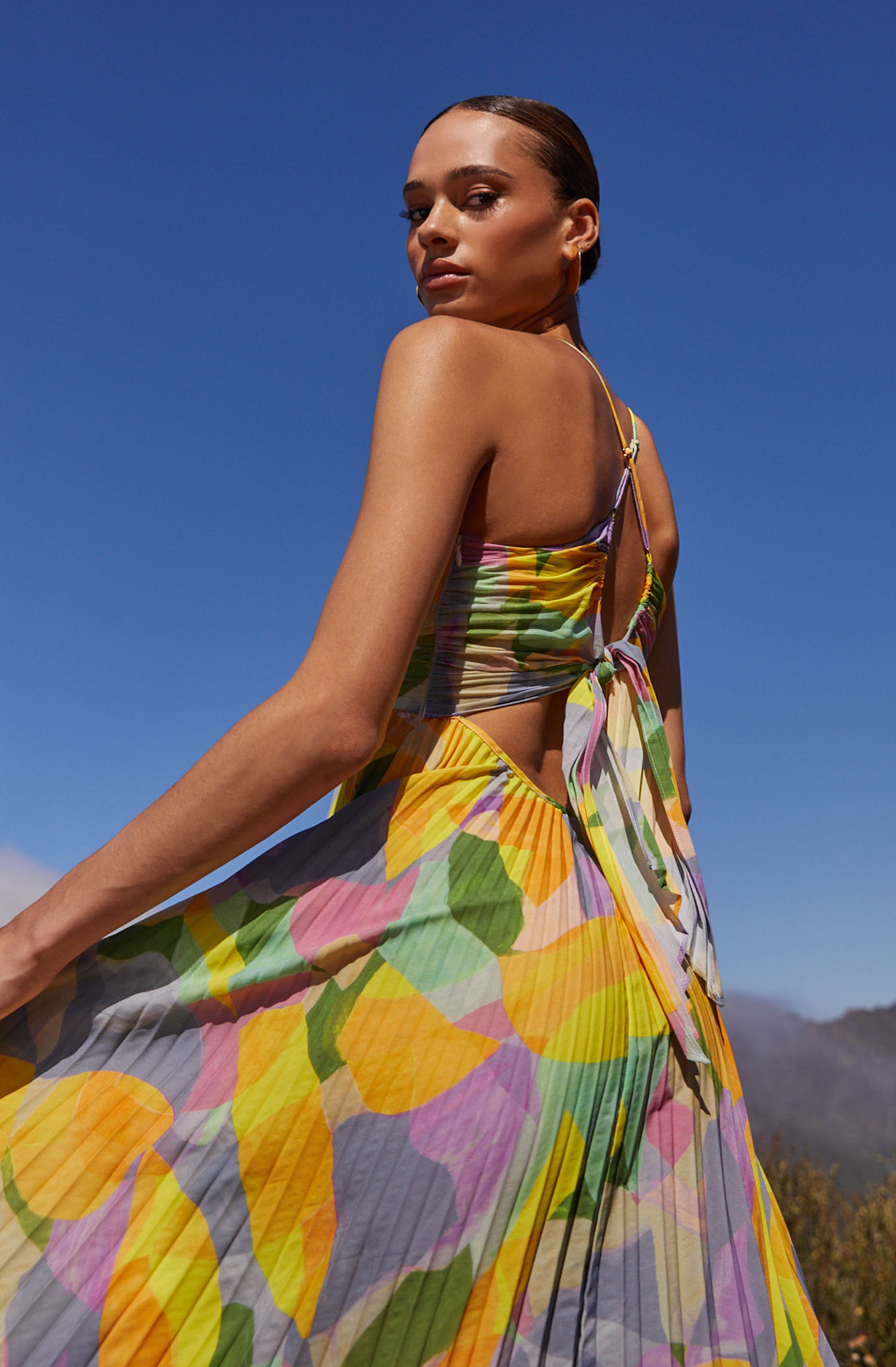 A woman is wearing a sleeveless, tie-up dress with a colorful, abstract floral print. The dress features a fitted bodice with spaghetti straps and a front tie, and a flowing pleated skirt. The pattern includes shades of yellow, pink, green, and purple, creating a vibrant and lively appearance.