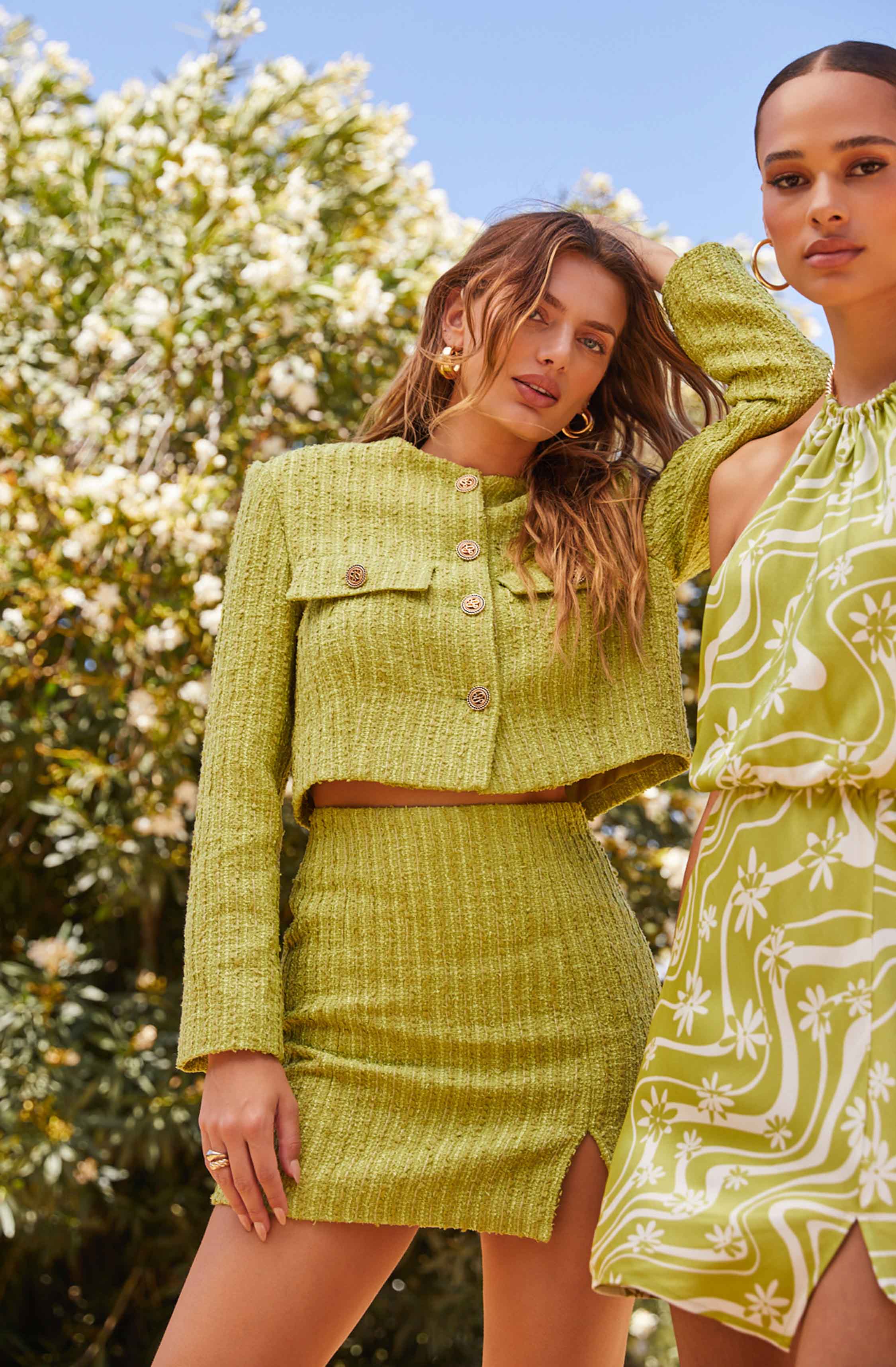 Two women are wearing coordinated lime green, textured outfits. One woman has on a cropped jacket with large buttons and a matching high-waisted skirt with a slit, while the other woman is dressed in a sleeveless, flowy dress with a white floral and geometric pattern. Both women are accessorized with hoop earrings and rings, and are outdoors with a background of blooming trees and a clear blue sky.